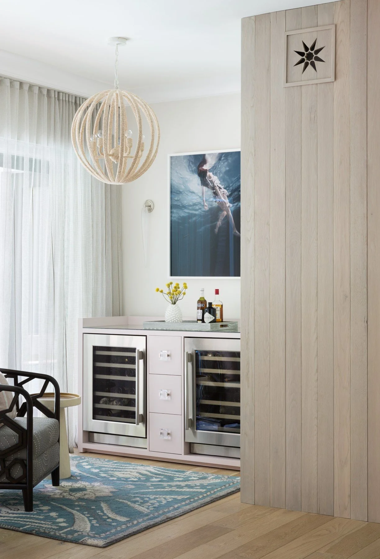 Interior of a modern living space with a white bar cabinet, wine bottles, a flower vase, a photo of an underwater swimmer, and a round hanging light fixture.