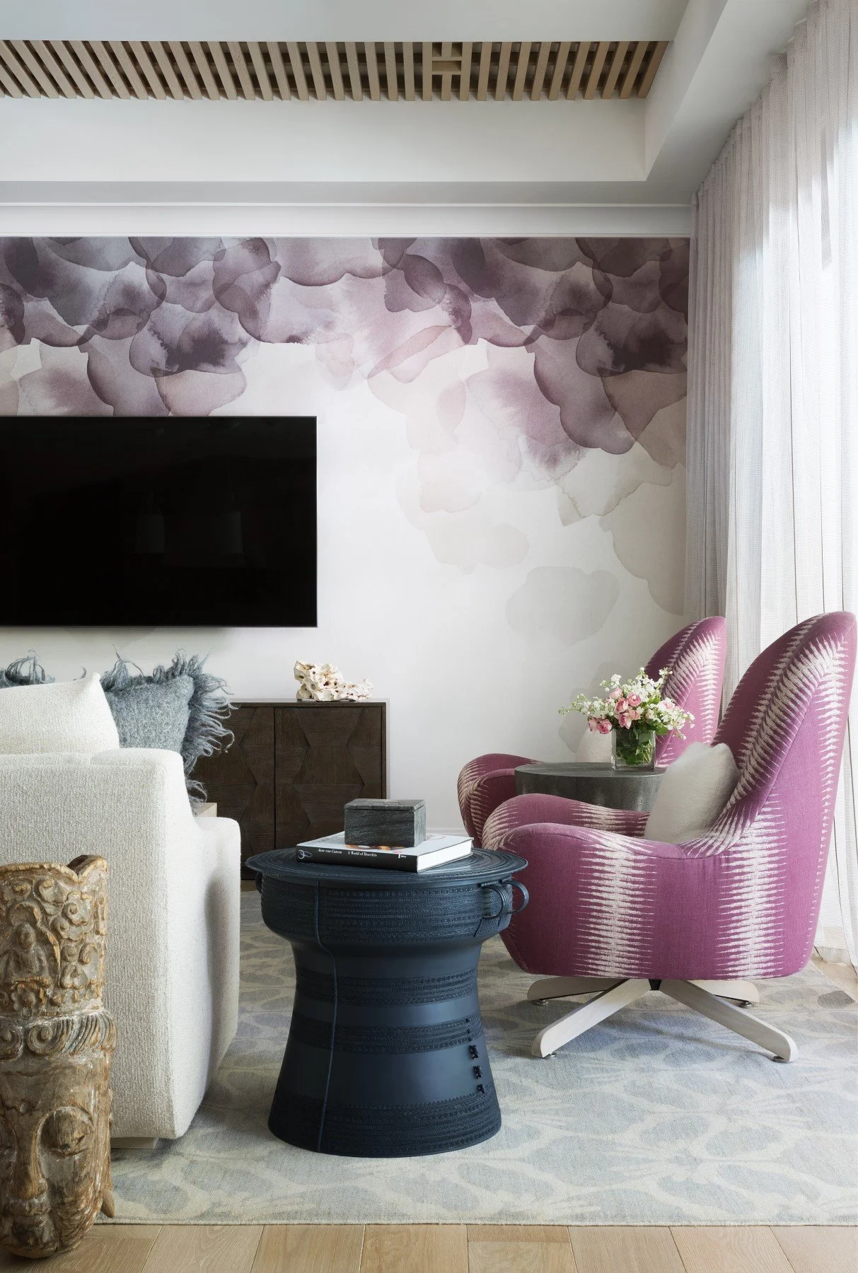 A living room with a wall-mounted flat screen TV, a dark wood cabinet, a white armchair, two pink patterned armchairs, and a black round coffee table. The room has a watercolor floral mural on the wall, a light-colored area rug, and sheer curtains le