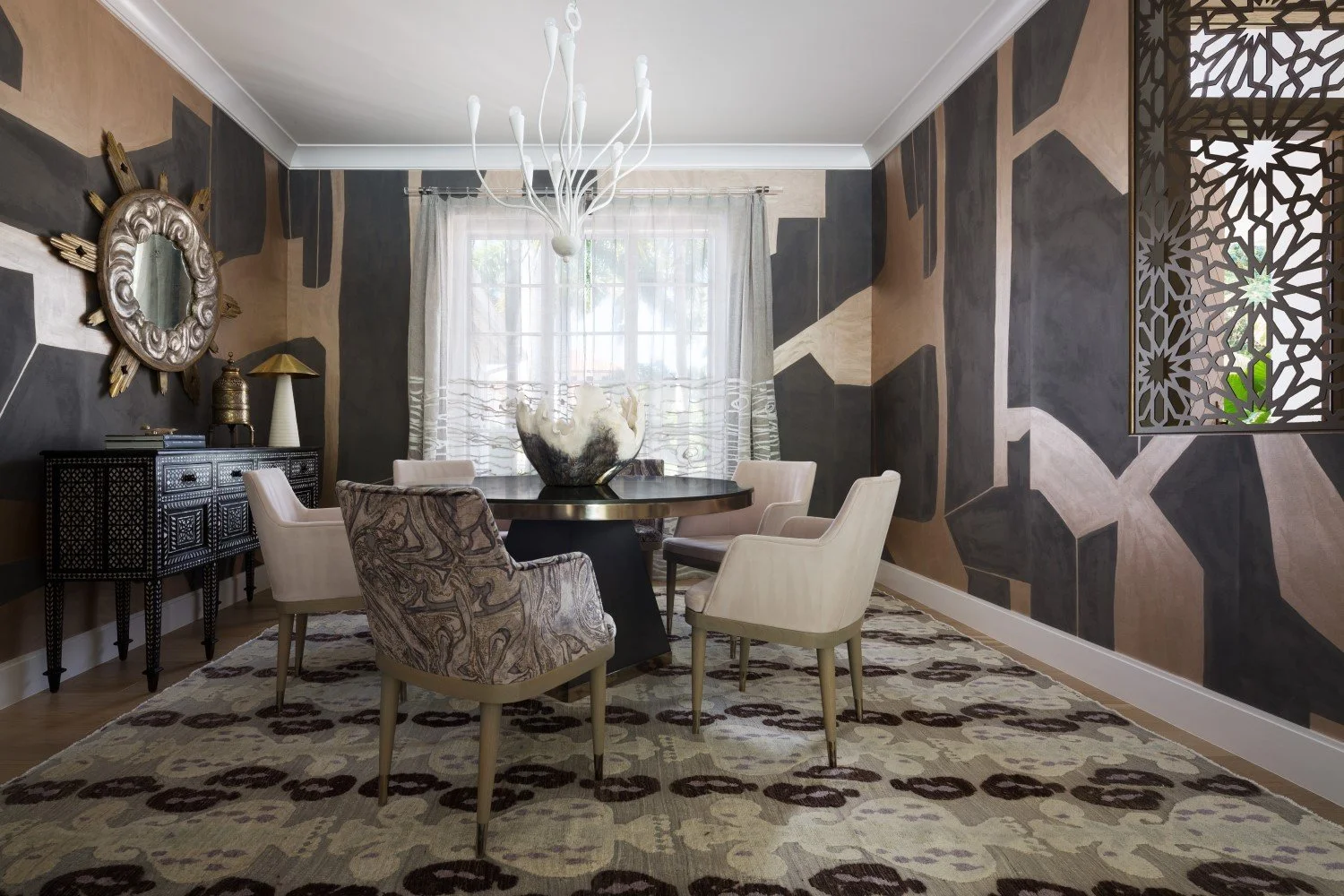 A dining room with geometric patterned walls, a window with semi-sheer curtains, and a chandelier. There is a round table with chairs, a decorative bowl, a sideboard, and a rug with a floral pattern.