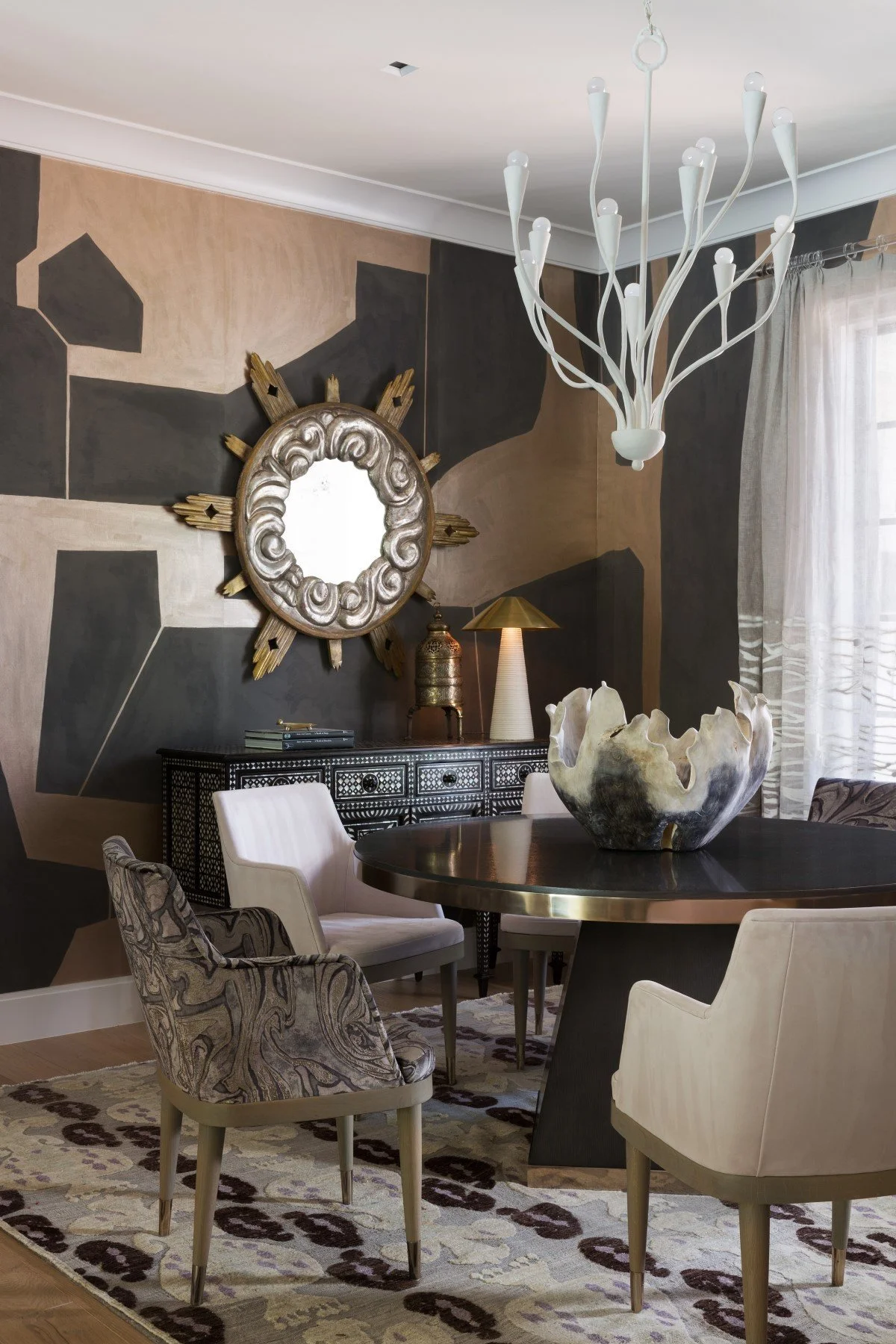Interior of a dining room with a black round table, four chairs, and a black sideboard. A decorative mirror with a sunburst frame hangs on a geometric-patterned wall. A modern white chandelier hangs from the ceiling, and a large decorative bowl sits 