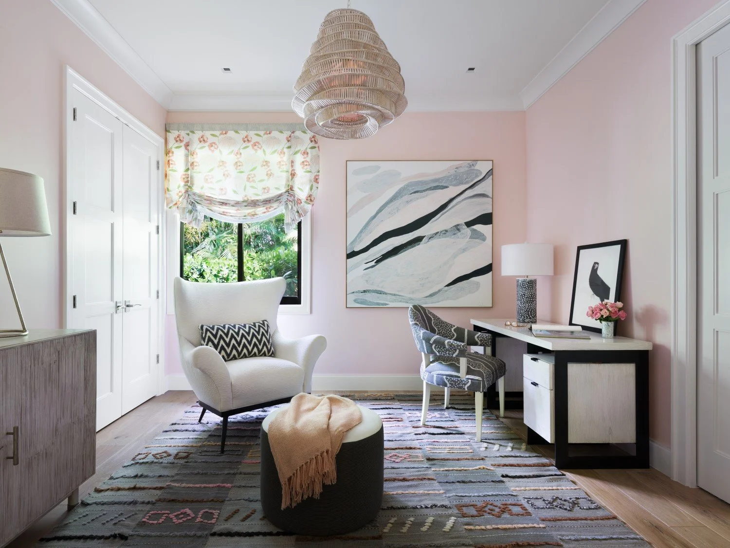 A cozy, bright room with pink walls, a window with floral valance, white armchair with black and white pillow, patterned desk and chair, abstract landscape painting, framed bird art, vase of flowers, and modern lighting fixtures.