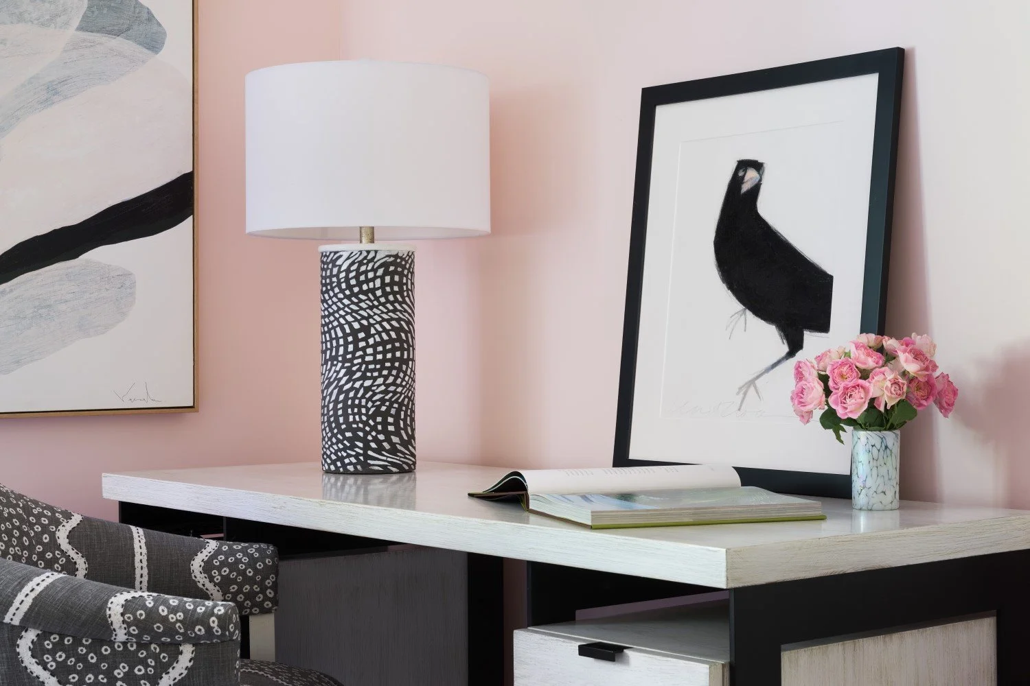 Decorative table with a black and white patterned lamp, a framed black bird illustration, a vase of pink roses, and an open book.