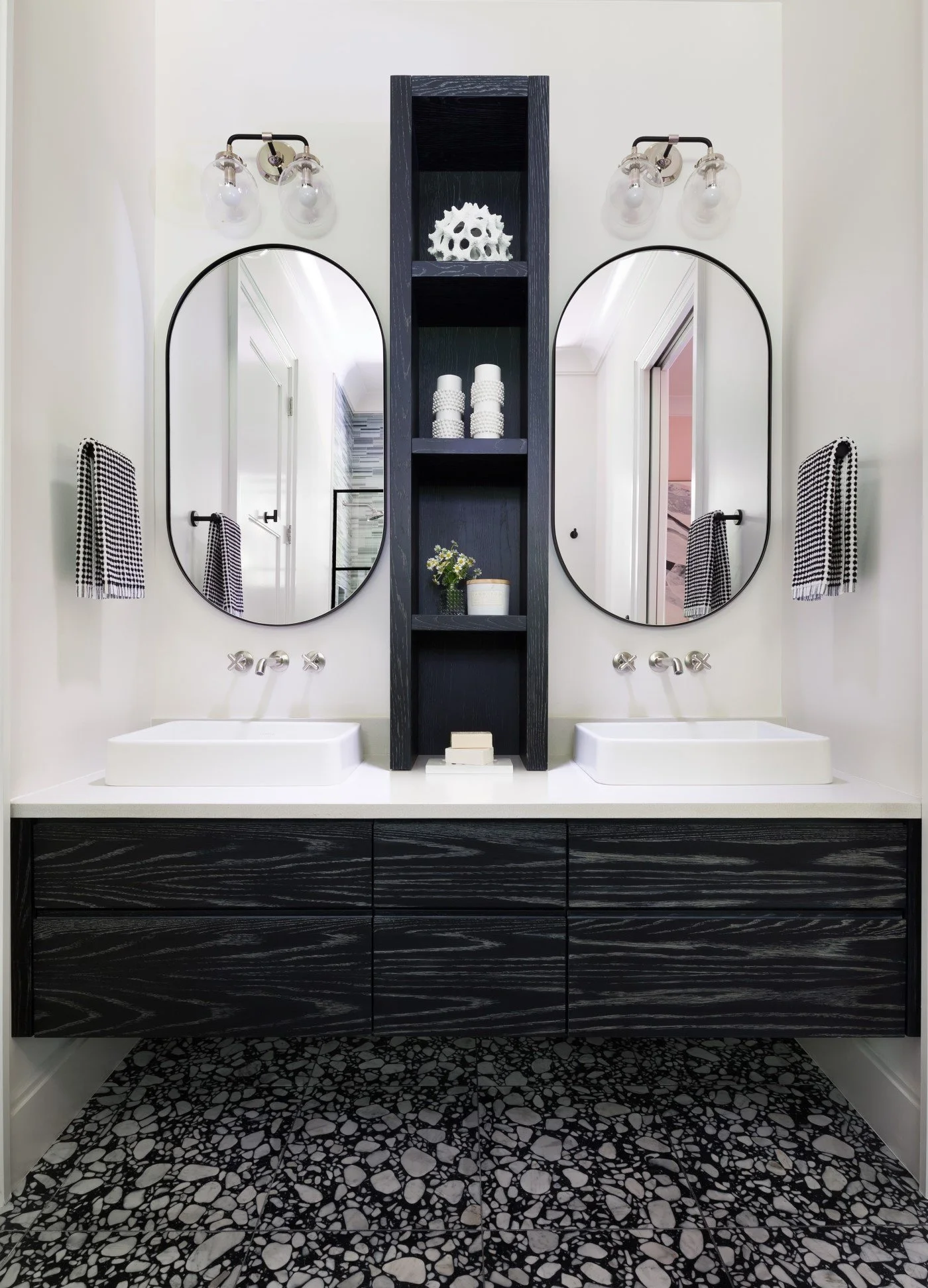 Modern bathroom with double vanity, black cabinetry, white sinks, oval mirrors, black and white patterned floor, wall-mounted light fixtures, towels, and decorative items on a dark shelf.