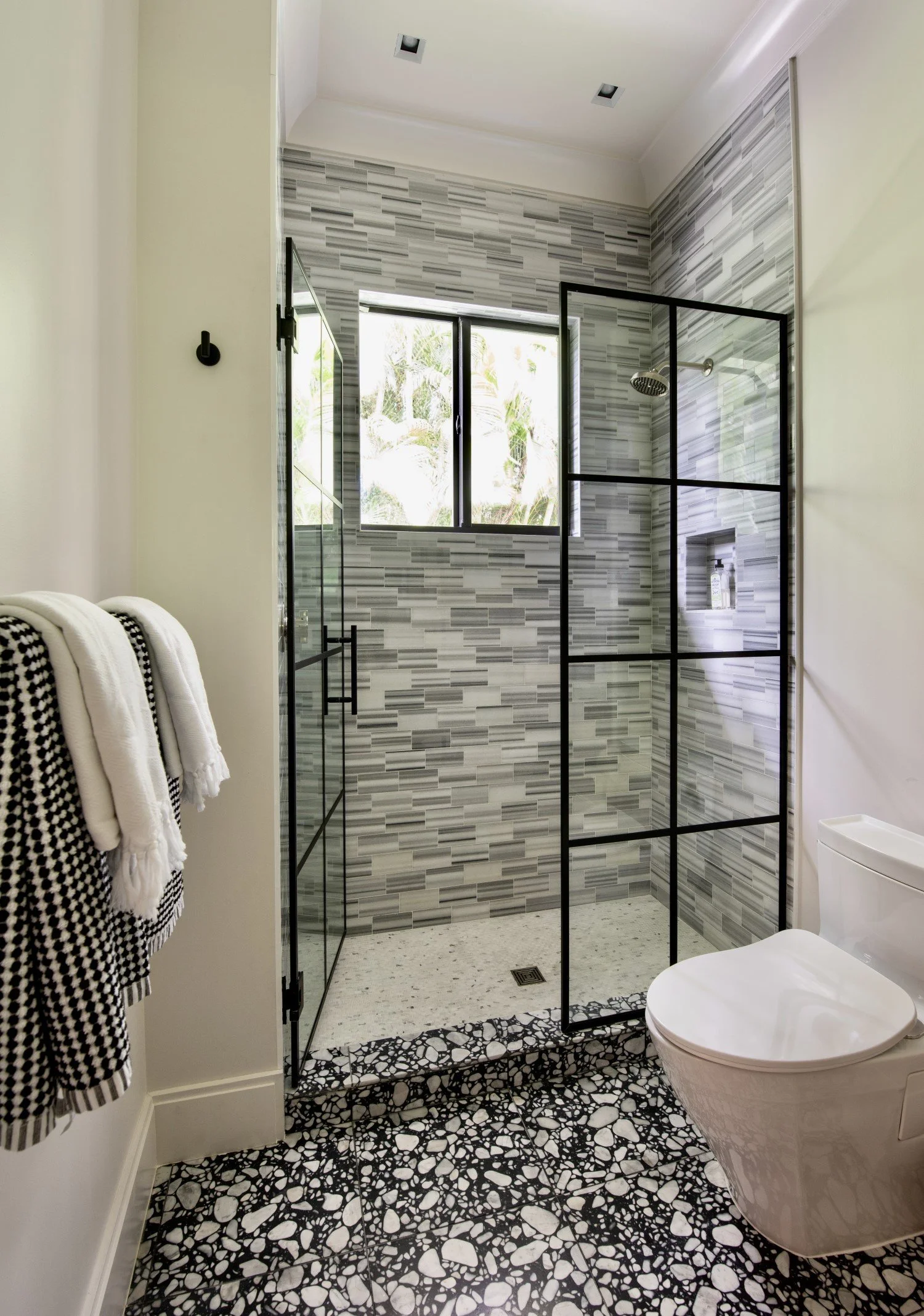 Modern bathroom with a glass shower enclosure, window, and black and white patterned terrazzo floors.