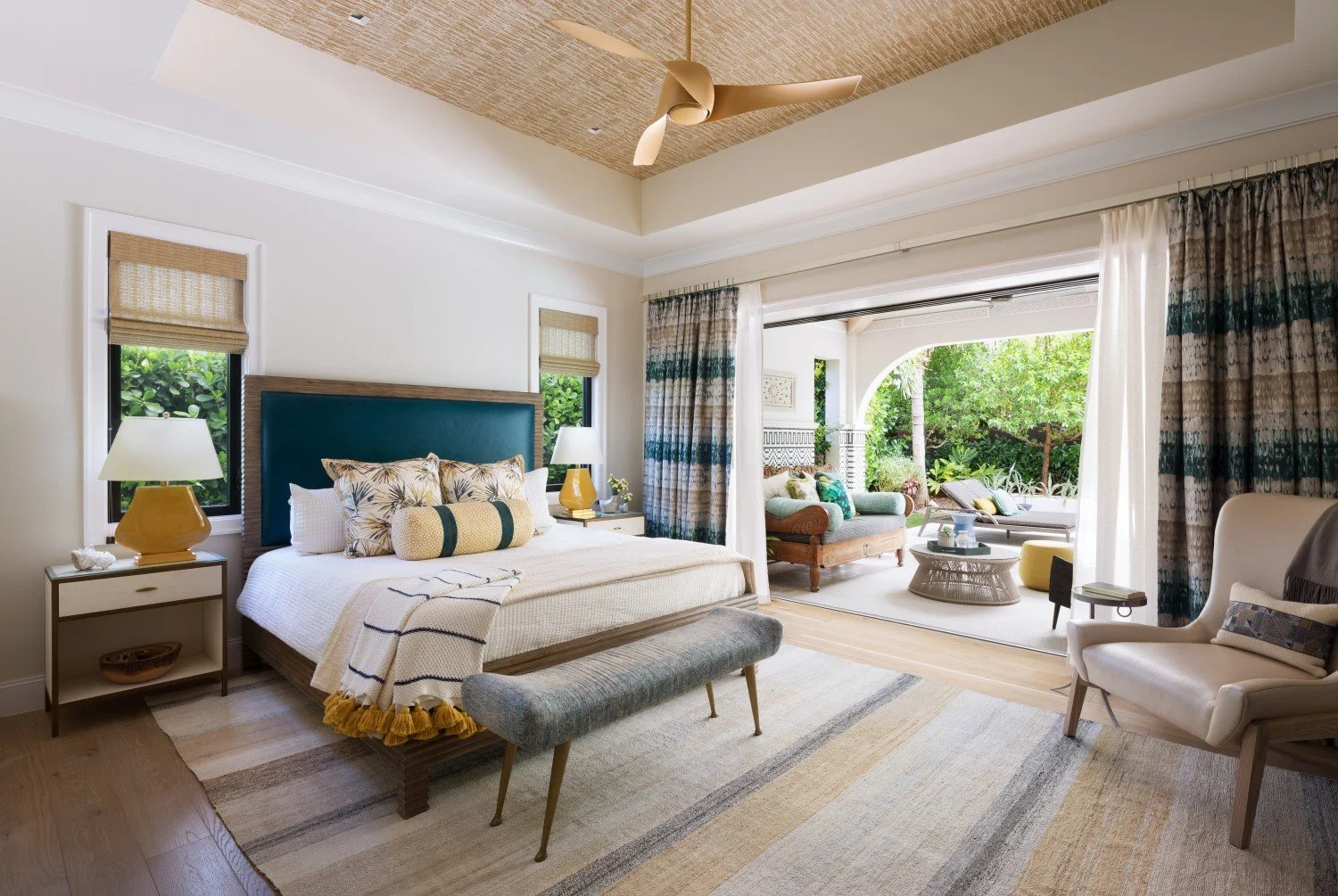 A bright, airy bedroom with a king-sized bed, nightstands with yellow lamps, and a sitting area outside on a patio with lush greenery.