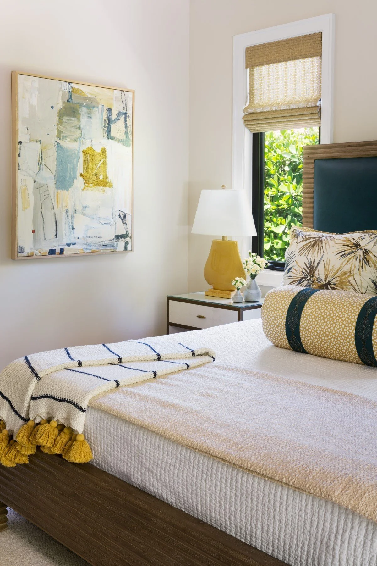 A cozy bedroom with a bed, decorative pillows, a white blanket with tassels, a yellow lamp, a nightstand with small vases and flowers, a window with checked blinds and greenery outside, and an abstract wall art.