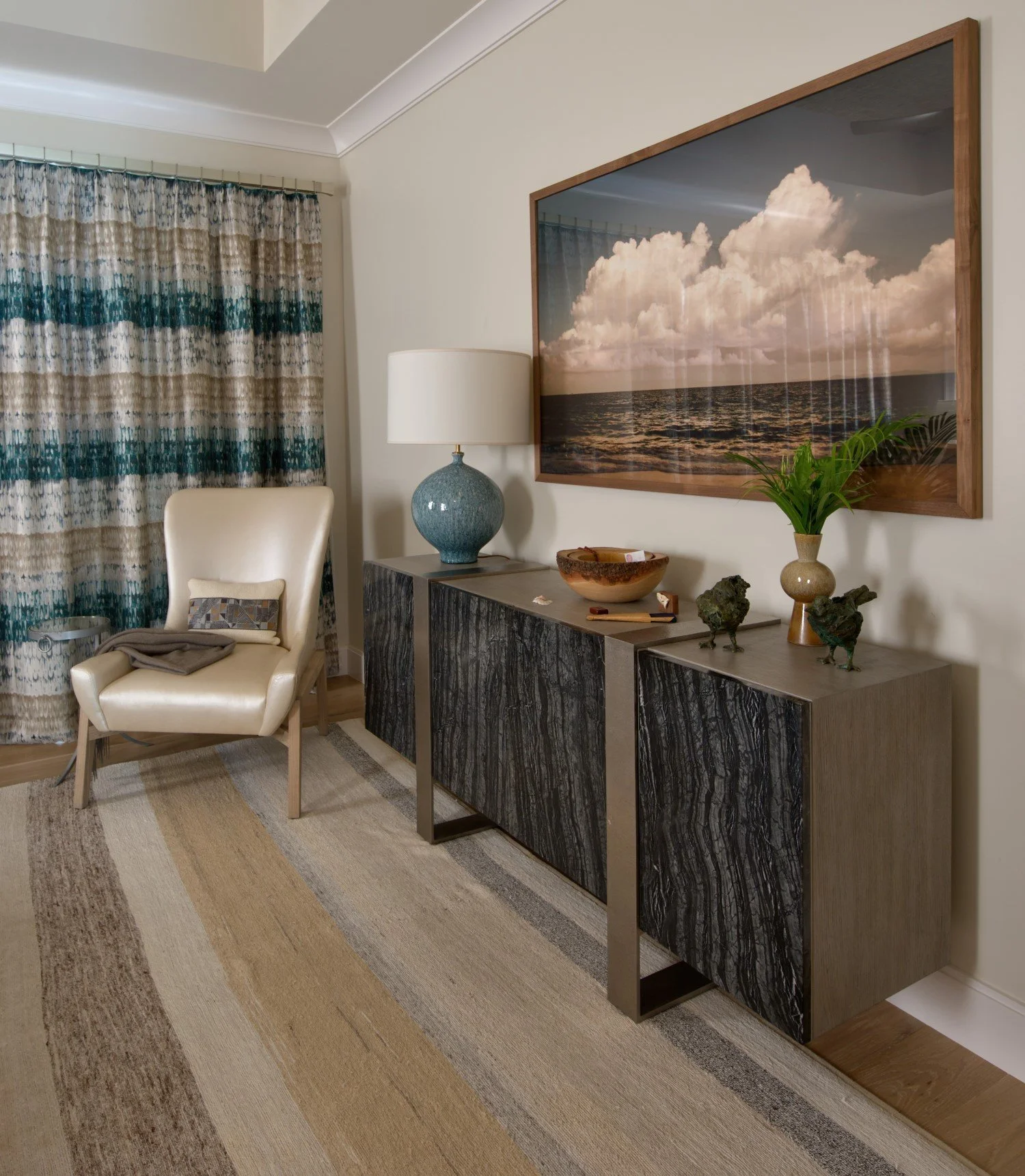 A living room corner with a white armchair, a wooden patterned sideboard, a lamp, a large framed photo of the ocean and sky, and decorative items including a vase with greenery and small sculptures.