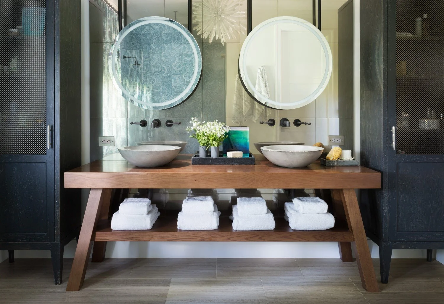 A modern bathroom vanity with two circular mirrors above two vessel sinks, a wooden countertop, and black wall-mounted faucets. Below the counter are neatly folded white towels. The vanity is flanked by black cabinets with mesh doors. On the counter 