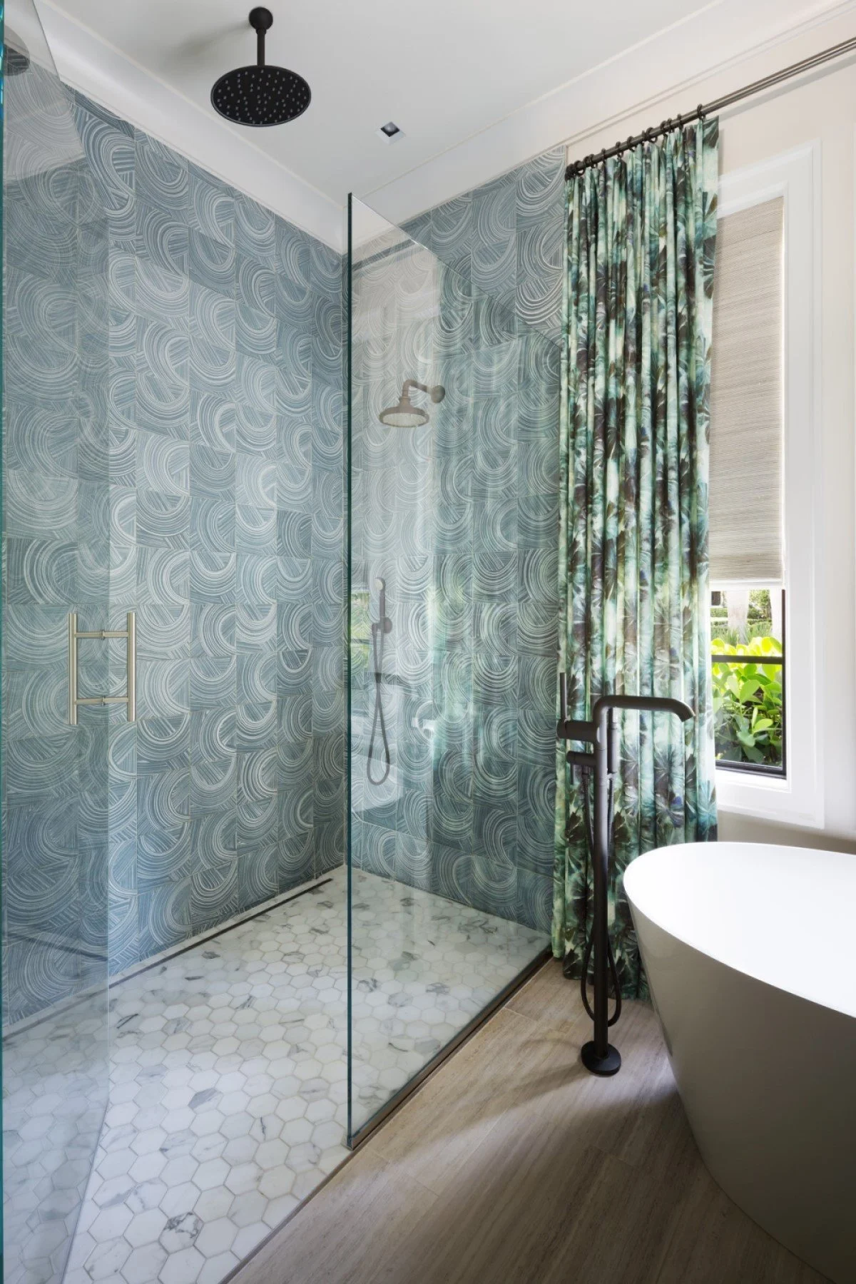 A modern bathroom with a glass-enclosed shower featuring blue patterned tiles, a rainfall showerhead, a window with floral curtains, a white freestanding bathtub, and a black toilet nearby.