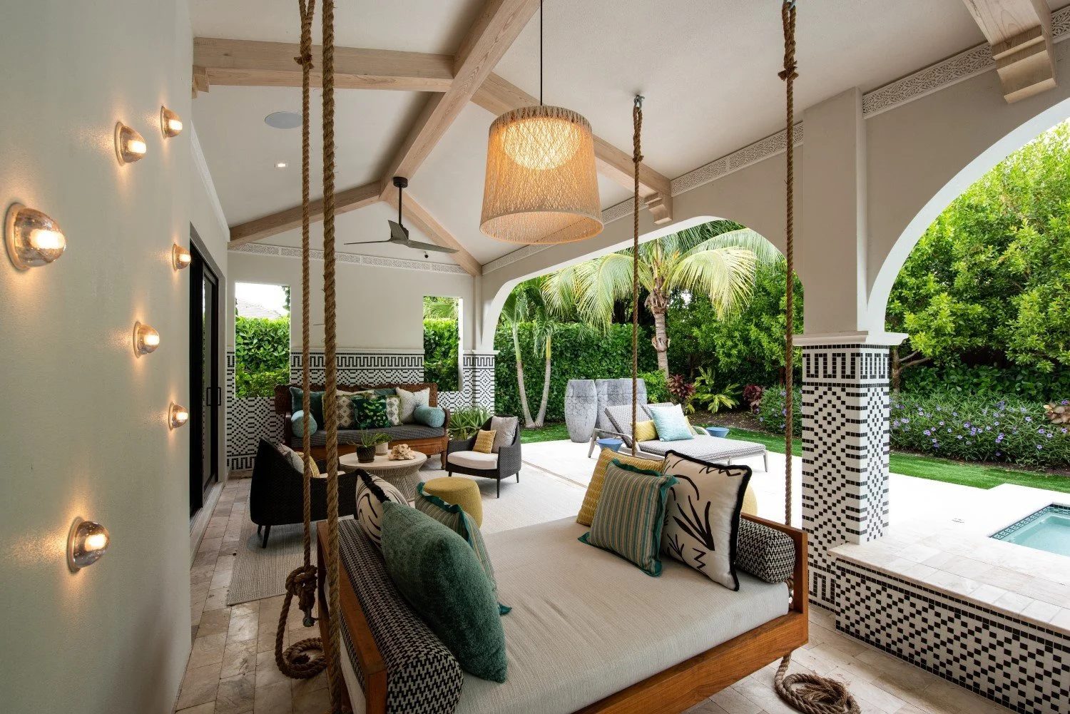 Outdoor patio with hanging bench swing, various seating options, lush greenery, and a small pool under a covered roof with wooden beams.