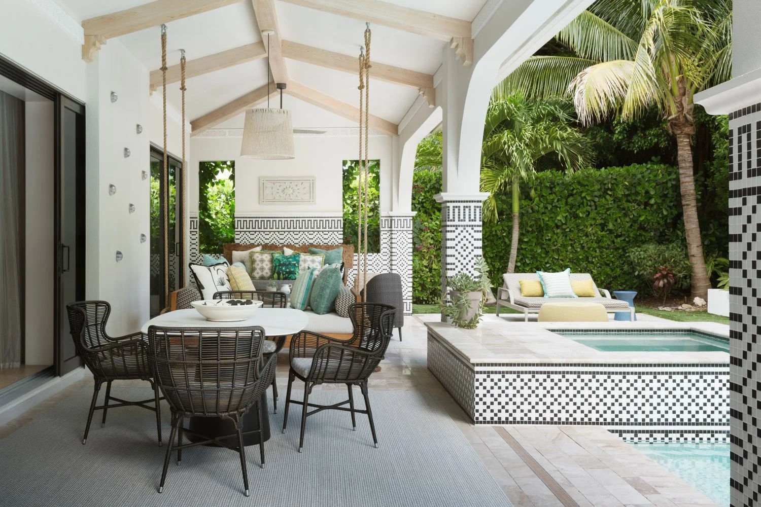 Outdoor patio with a dining table and chairs, a cushioned lounge area, and a hot tub with a mosaic tile pattern, surrounded by lush greenery and palm trees.