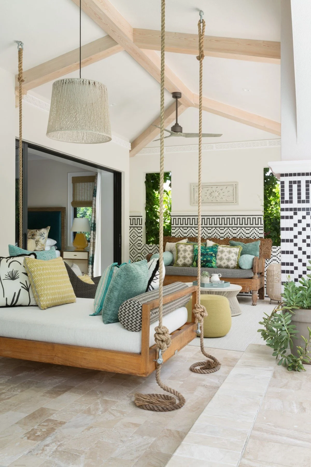 Living room with a wooden swing seat hanging from the ceiling, decorated with various cushions, a white coffee table, and an indoor plant. Background features a wall with black-and-white patterned tiles, a wooden bench with cushions, and windows with