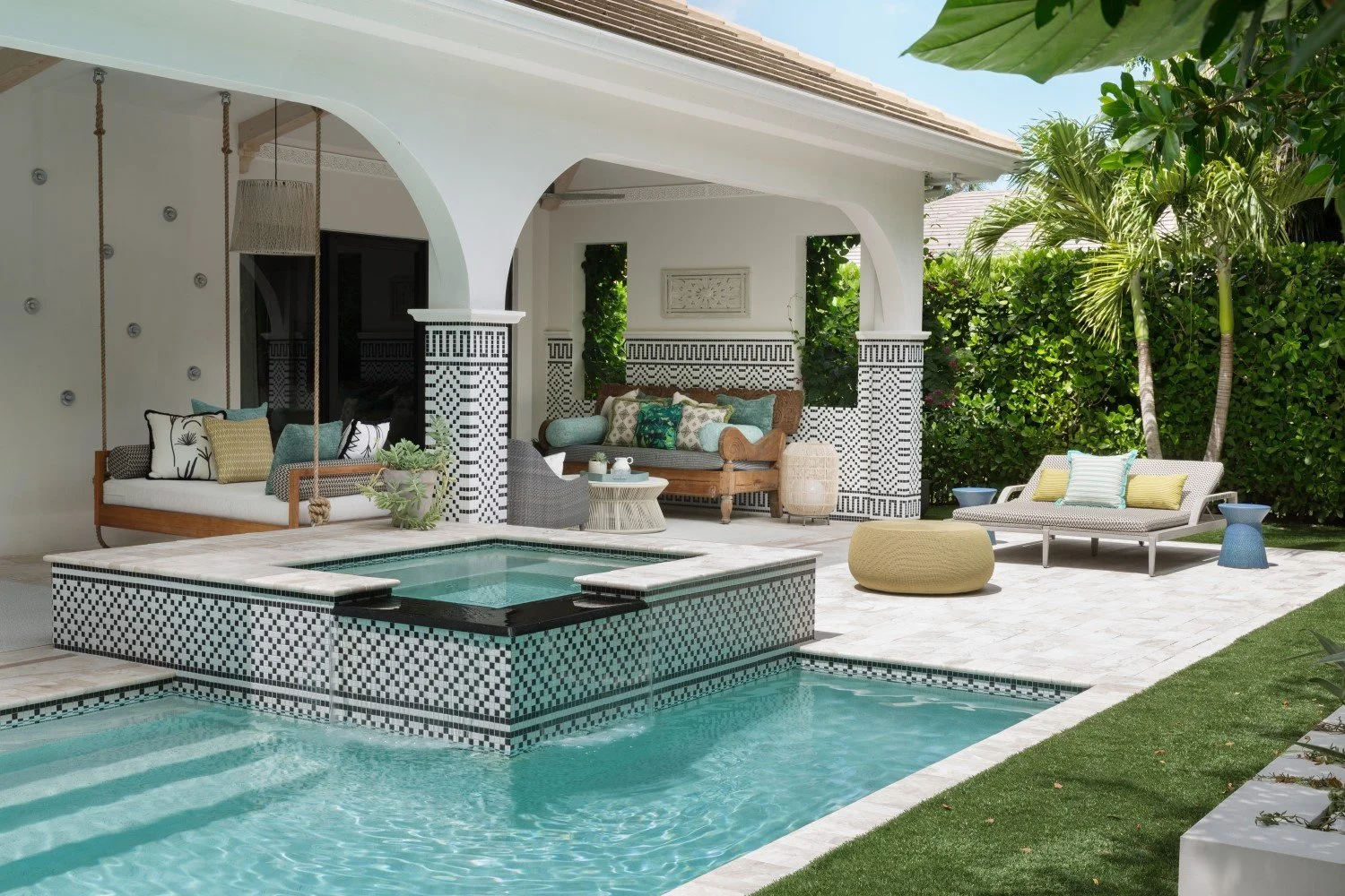 Outdoor patio with a swimming pool featuring a raised hot tub with black and white mosaic tiles, multiple lounge chairs with colorful pillows, tropical plants, and a white house with architectural arches and black and white decorative tiles.