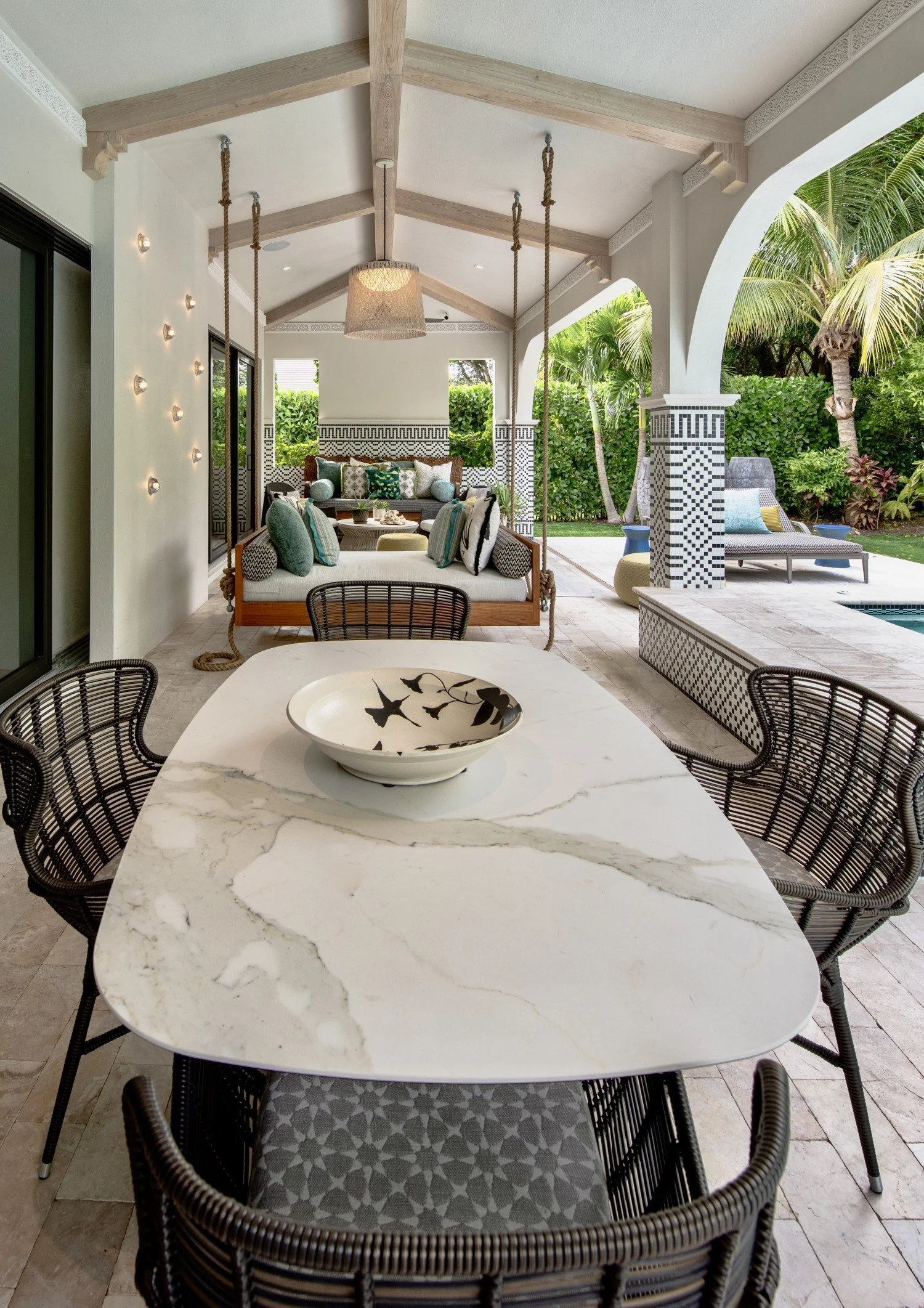 Outdoor covered patio with dining table, wicker chairs, decorative bowl, hanging swing, cushioned seating area with throw pillows, poolside lounge chair, lush greenery, and tiled columns.