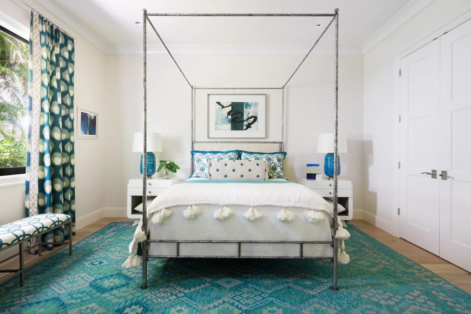 Bedroom with a white bed, decorative pillows, and a canopy frame, flanked by two white nightstands with blue lamps, window with patterned curtains, and a green and blue area rug.