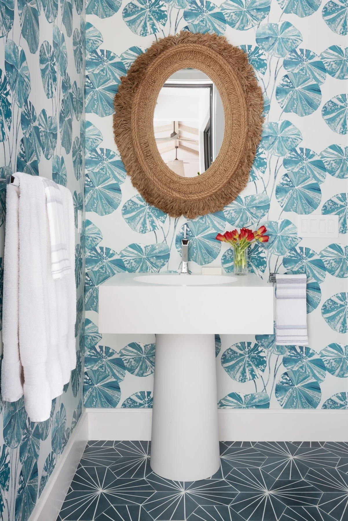 A bathroom with wallpaper featuring large blue and white leaf patterns, a white pedestal sink, a round mirror with a fringed wicker frame, a glass vase with red and yellow flowers, a white towel hanging on a silver towel bar, and black floor tiles wi