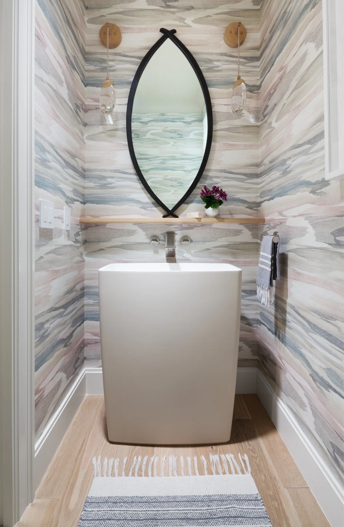 Small bathroom with swirling gray, pink, and white marble walls, a white rectangular sink, a large black-framed oval mirror, two hanging glass light fixtures, a wooden shelf with a potted purple flower, a towel bar with a gray and white towel, and a 