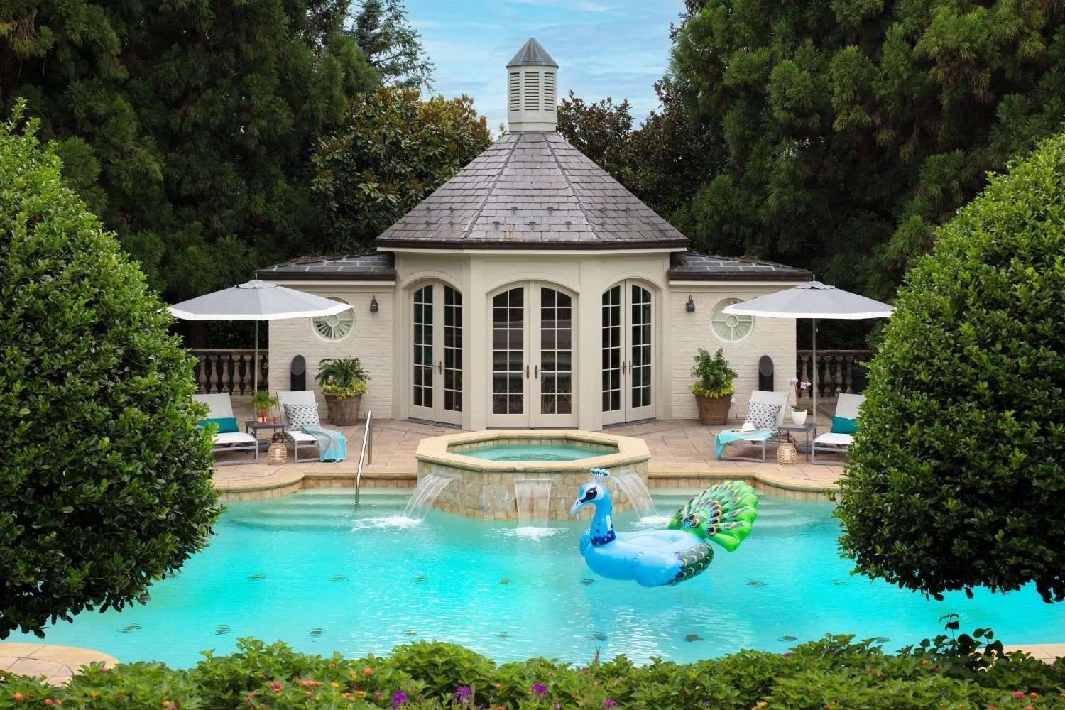 A backyard with a swimming pool featuring a blue peacock-shaped float, surrounded by two trees and poolside furniture including two umbrellas, lounge chairs, and potted plants in front of a small house with white brick walls and a shingled roof.
