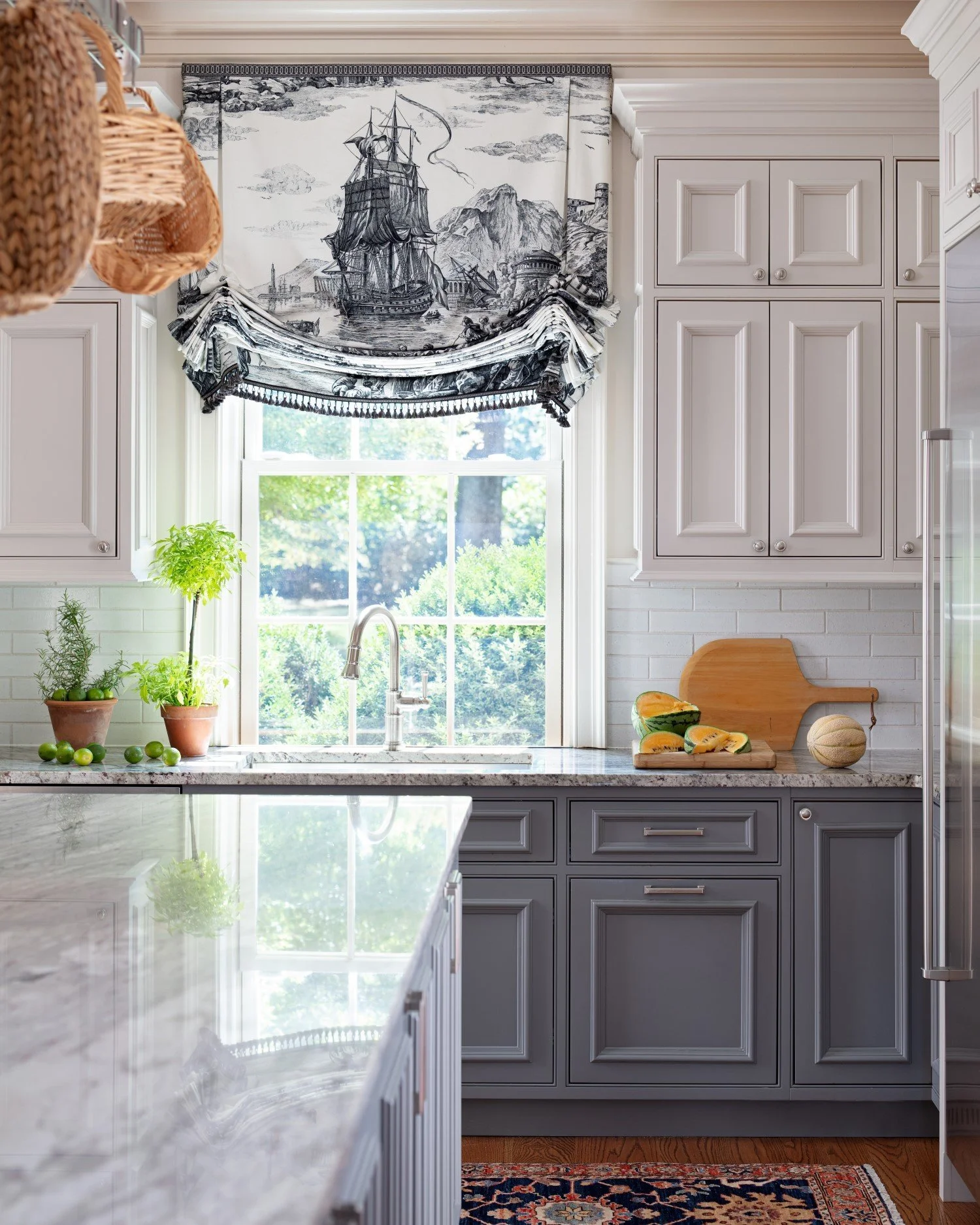 Kitchen with gray cabinets, a window with a nautical-themed valance, potted herbs, a cutting board with sliced melon, and a patterned rug.