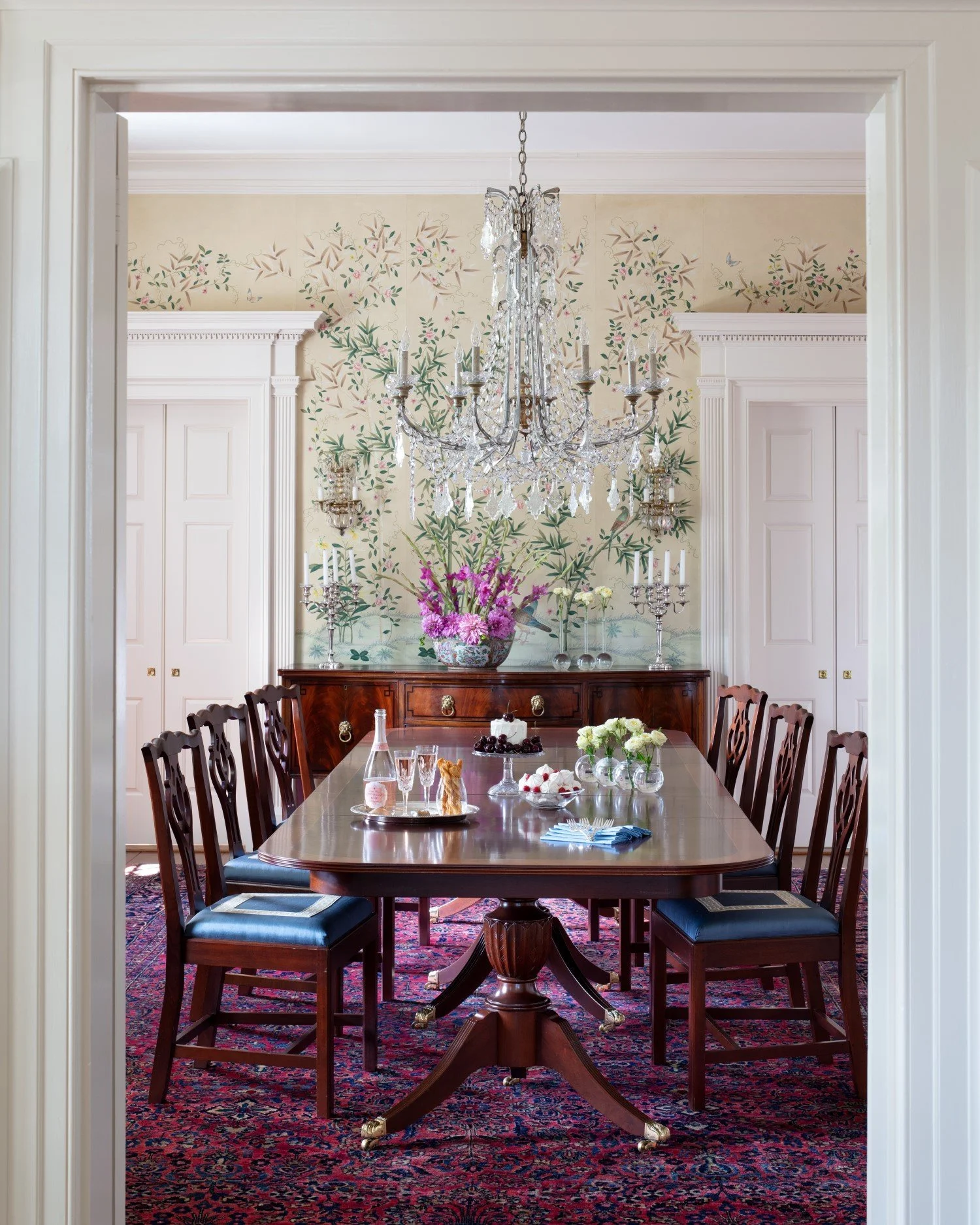 A formal dining room with a large wooden table, eight chairs with cushioned seats, a chandelier hanging from the ceiling, floral wallpaper, a sideboard decorated with flowers and candlesticks, and a rug underneath the table.