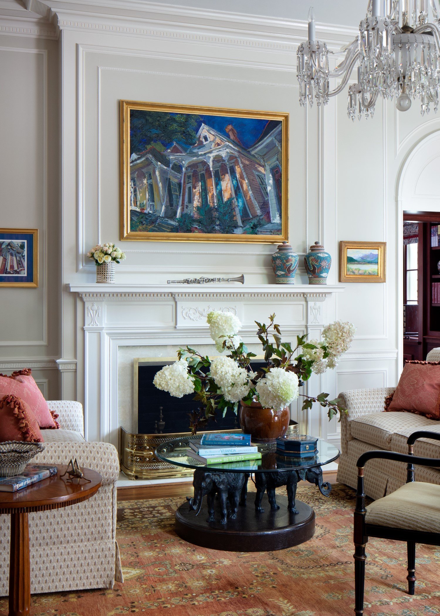 Living room with white walls and detailed molding, a fireplace mantel with decorative vases, a large framed painting of a house, smaller framed artworks, a chandelier, and a coffee table with books and a flower arrangement.