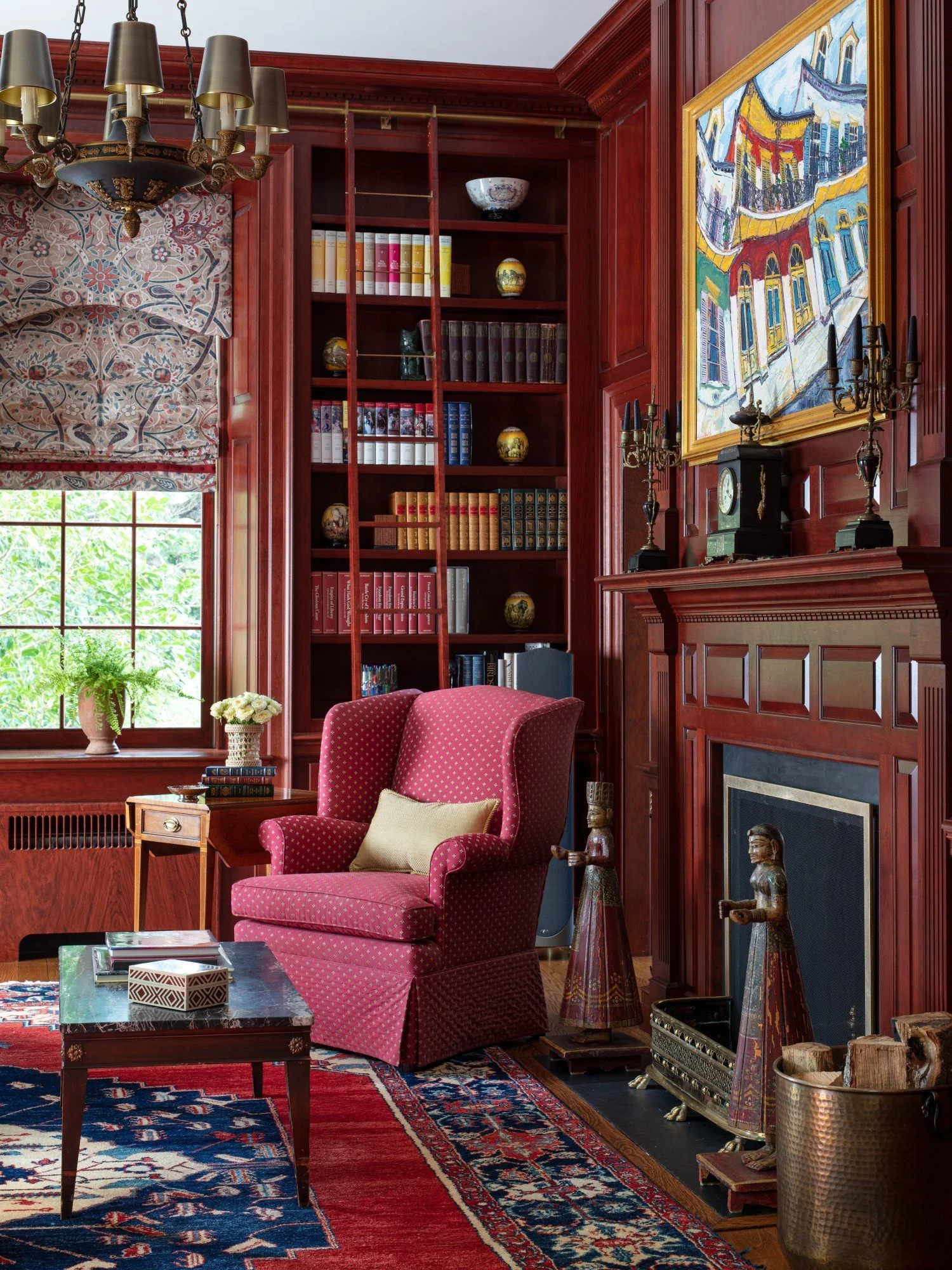 A cozy reading nook in a wood-paneled room featuring a pink upholstered armchair with a cream-colored pillow, a wooden side table with a potted plant and candles, a patterned rug, a dark fireplace with shelves above, a large colorful artwork, a windo