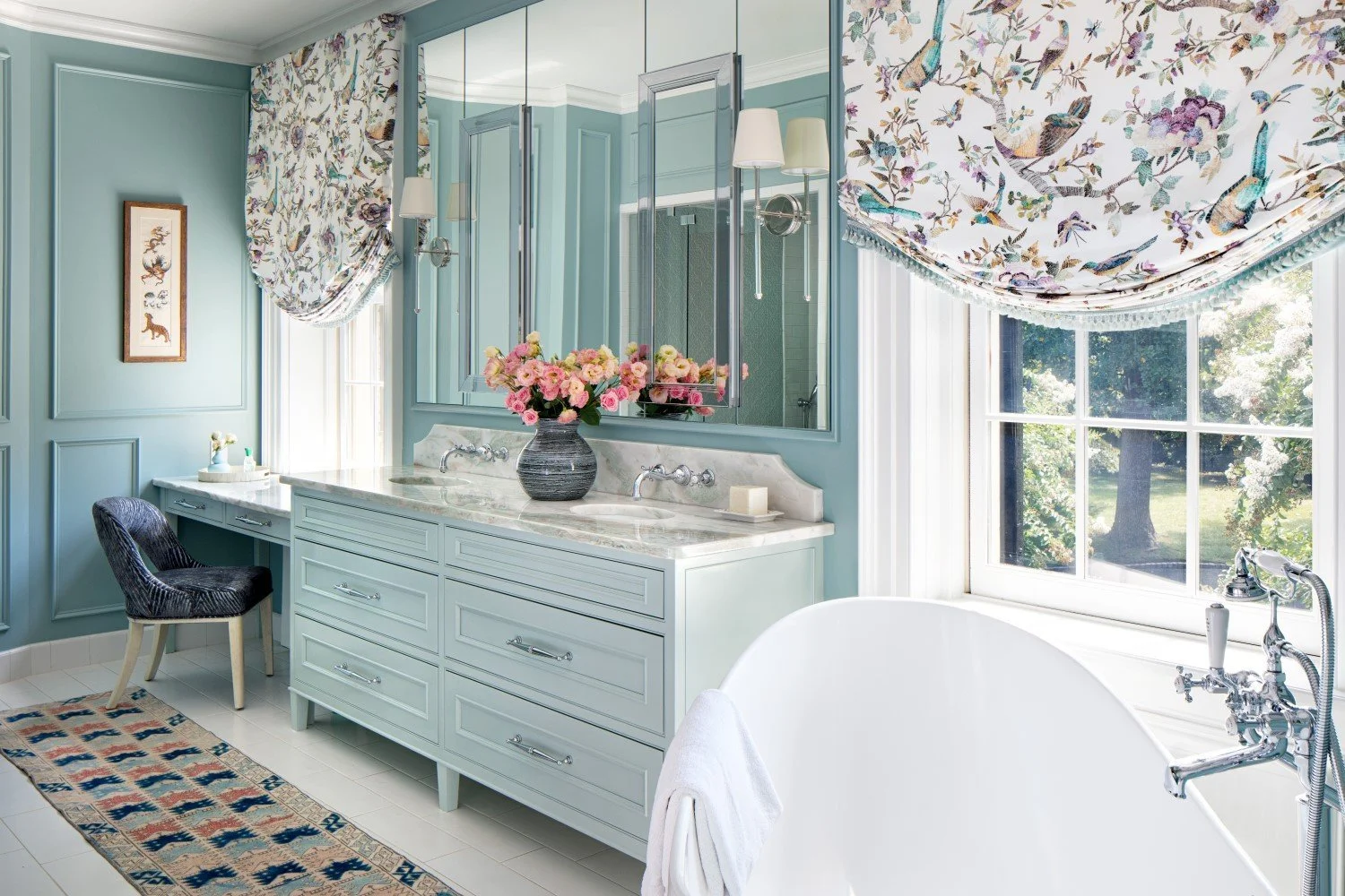 A bright, elegant bathroom with a large window, floral window treatments, light blue walls, a vanity with a marble countertop, a vase of pink flowers, a black chair, and a clawfoot bathtub with a white towel inside.