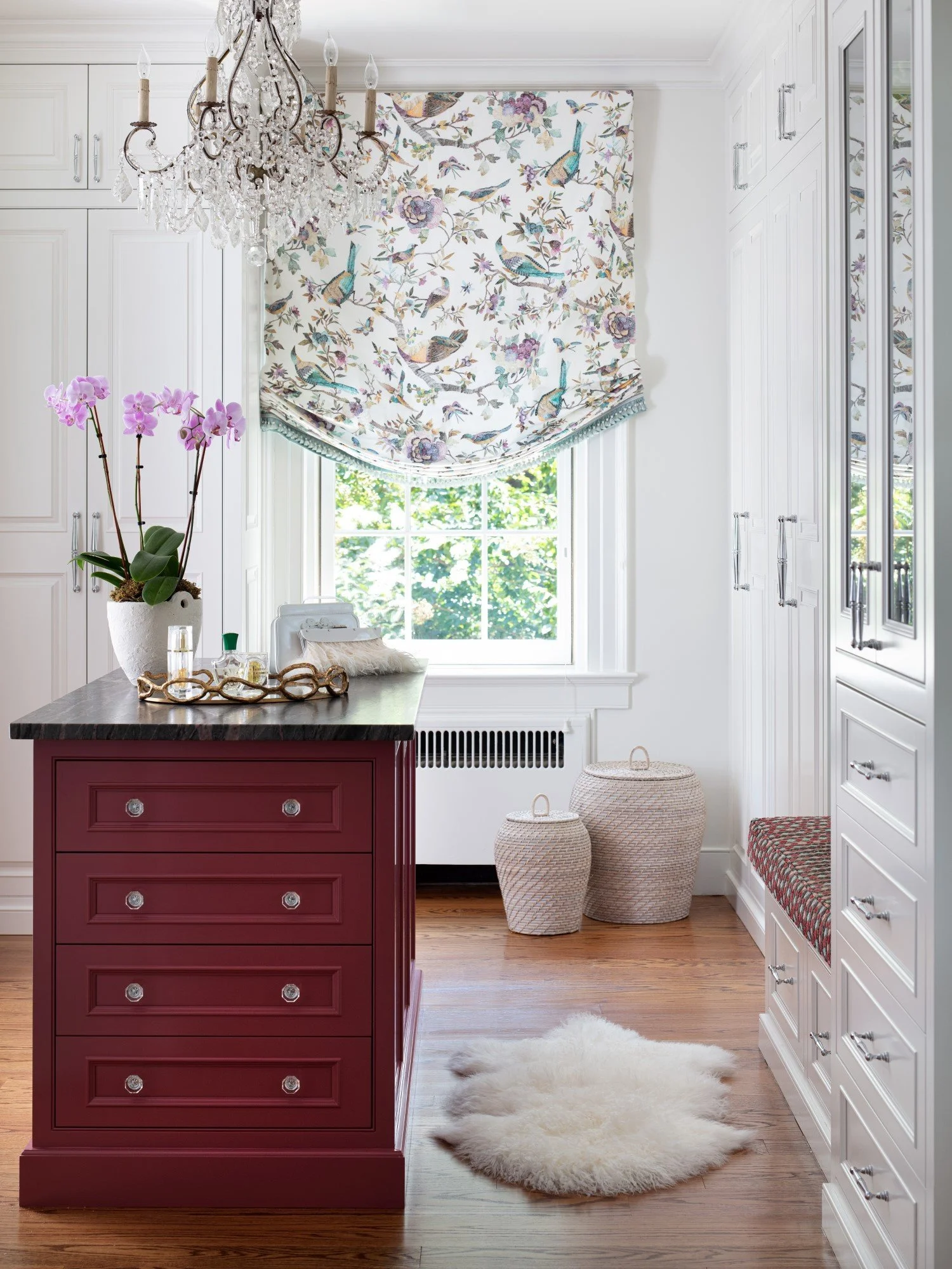A bright kitchen with white cabinets, a red kitchen island with a black marble top, a chandelier, and a window with a bird and floral patterned valance. There are purple orchids in a white pot, two woven baskets, a sheepskin rug, and a bench with a c