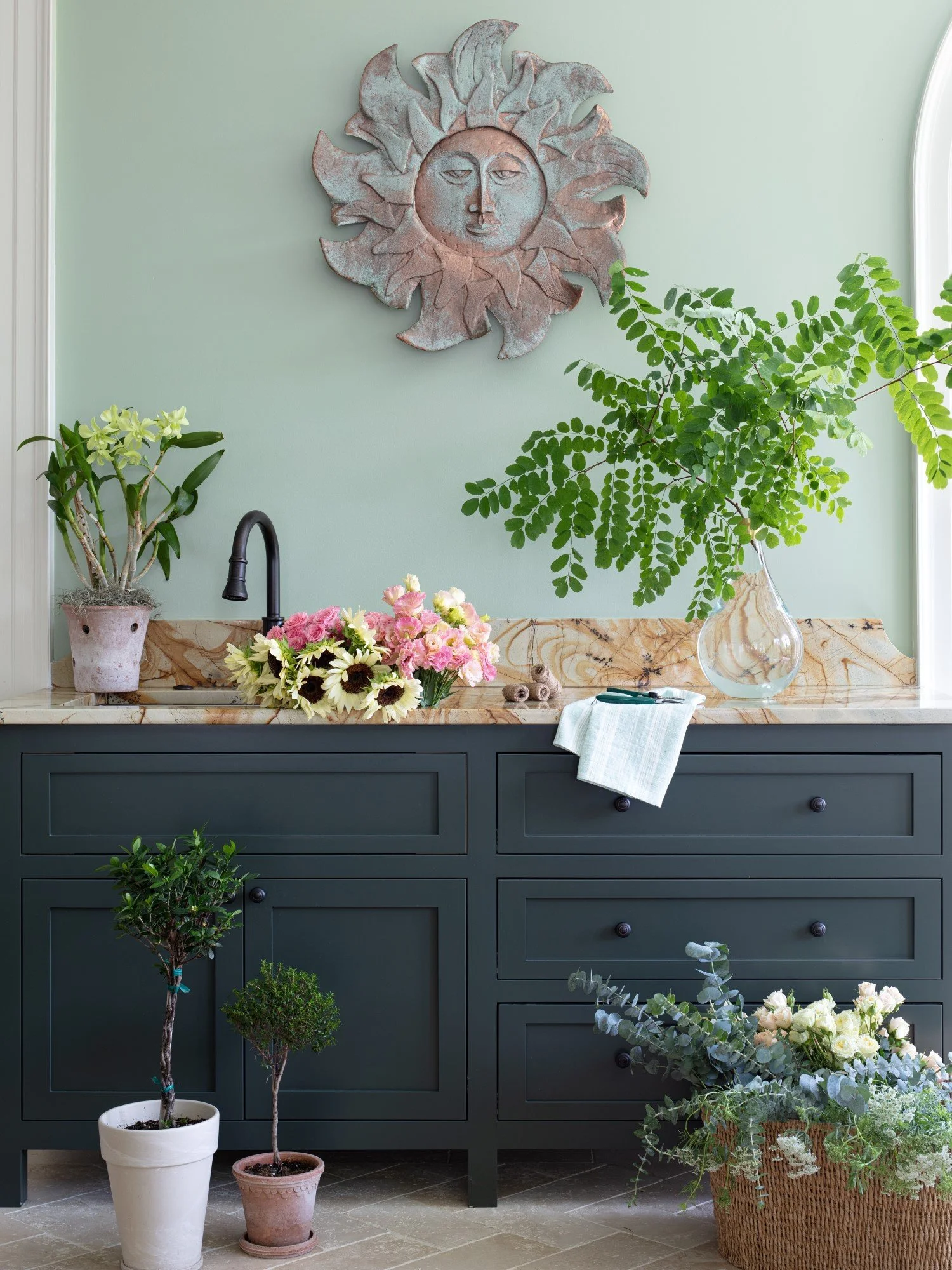 A decorated kitchen or utility space with a black cabinet, a marble countertop, a black faucet, and various plants, including potted and in a basket, with a sun face metal wall art above.