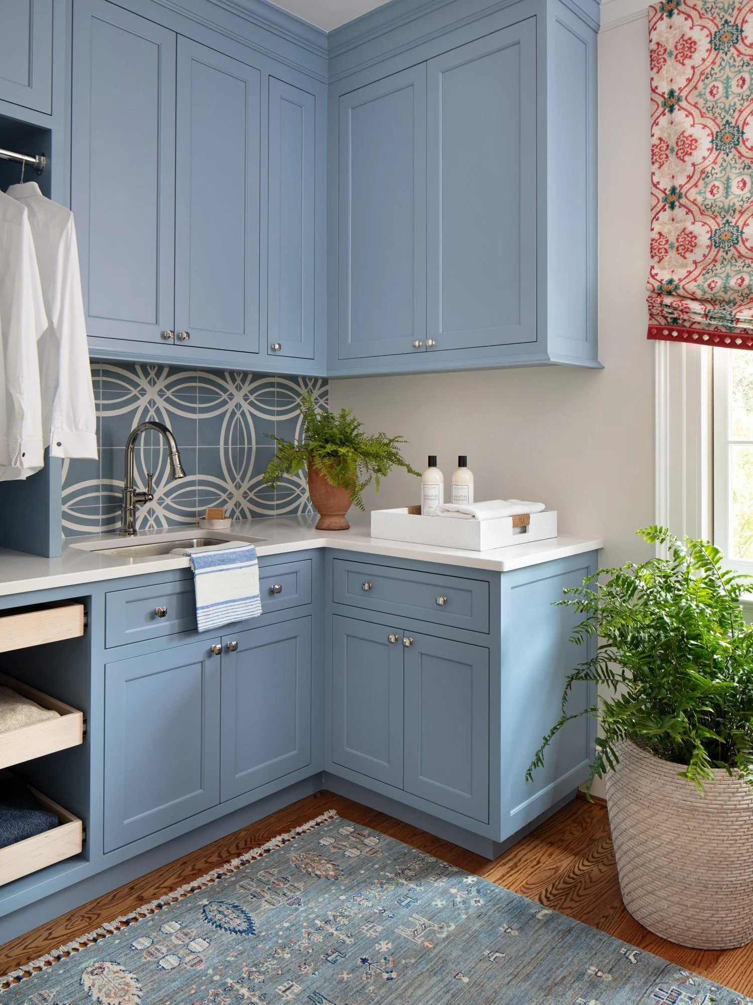 Corner of a cozy, well-organized laundry or utility room with blue cabinetry, a small sink, and decorative backsplash tile with geometric pattern. A cloth hangs on a drawer, and there are potted plants on the counter and floor, along with bottles and