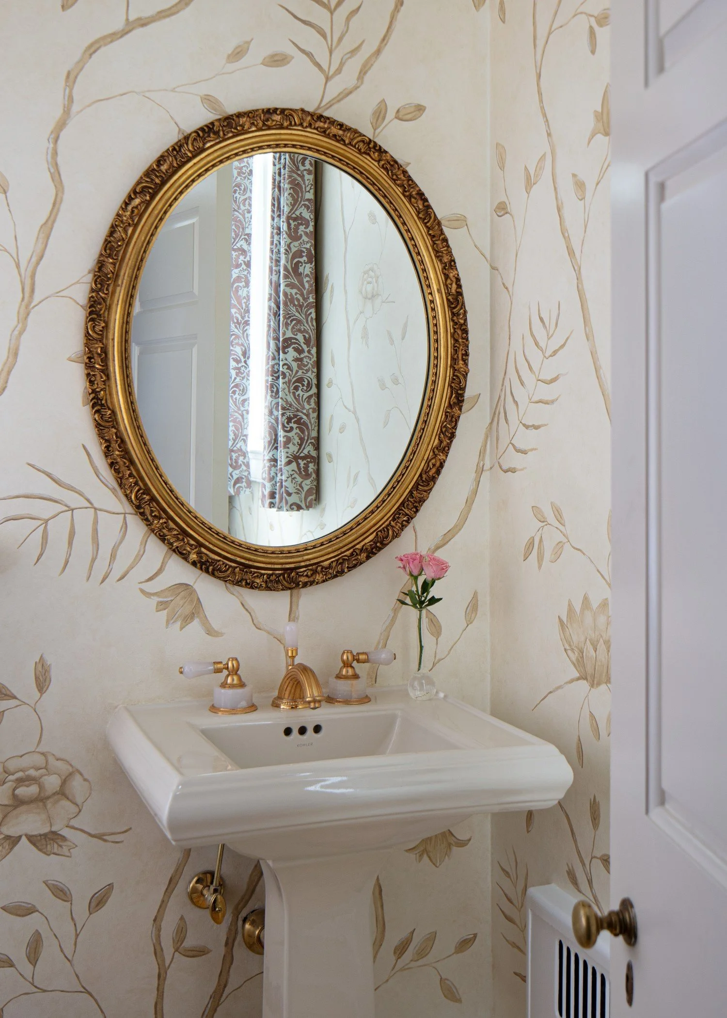 A vintage-style bathroom sink with a gold-trimmed oval mirror, two brass faucets, and a small vase with pink flowers on the sink ledge.