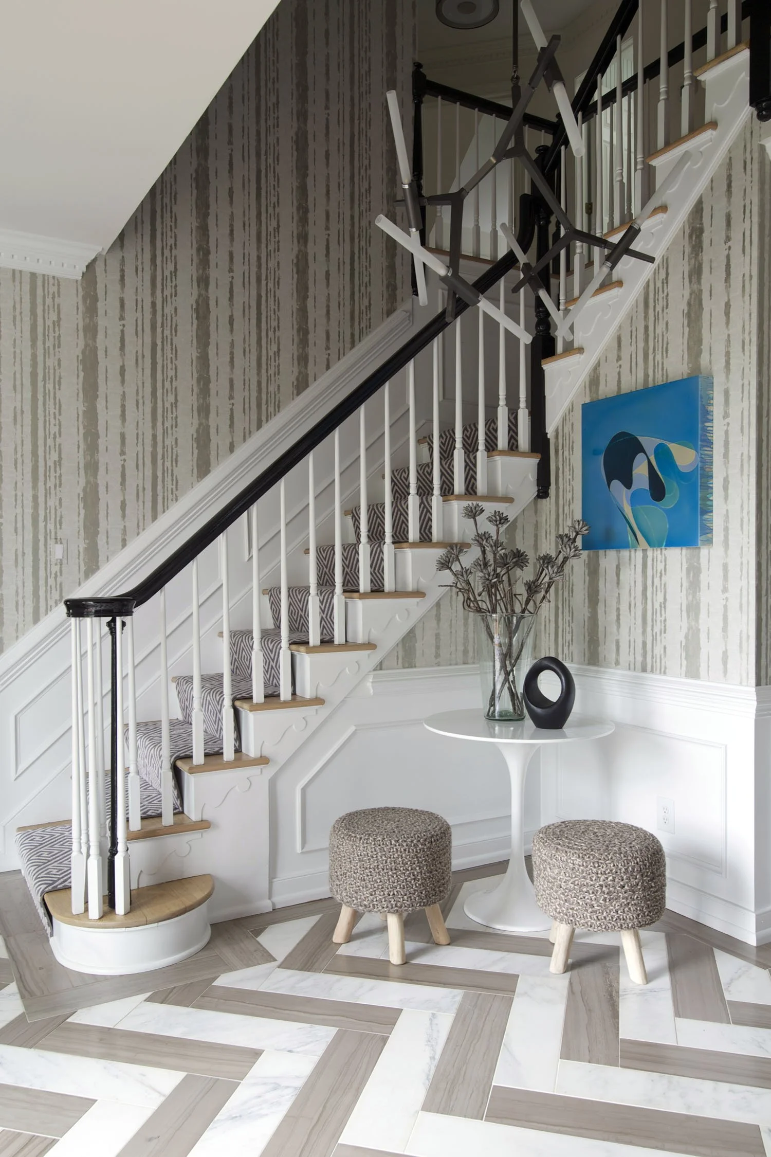 Interior of a house with a staircase, modern chandelier, abstract wall art, and a small sitting area with two stools and a round table.