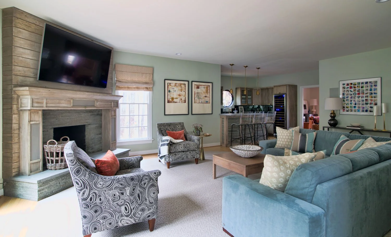 Living room with blue sectional sofa, patterned armchair, fireplace with large TV overhead, and kitchen in the background.