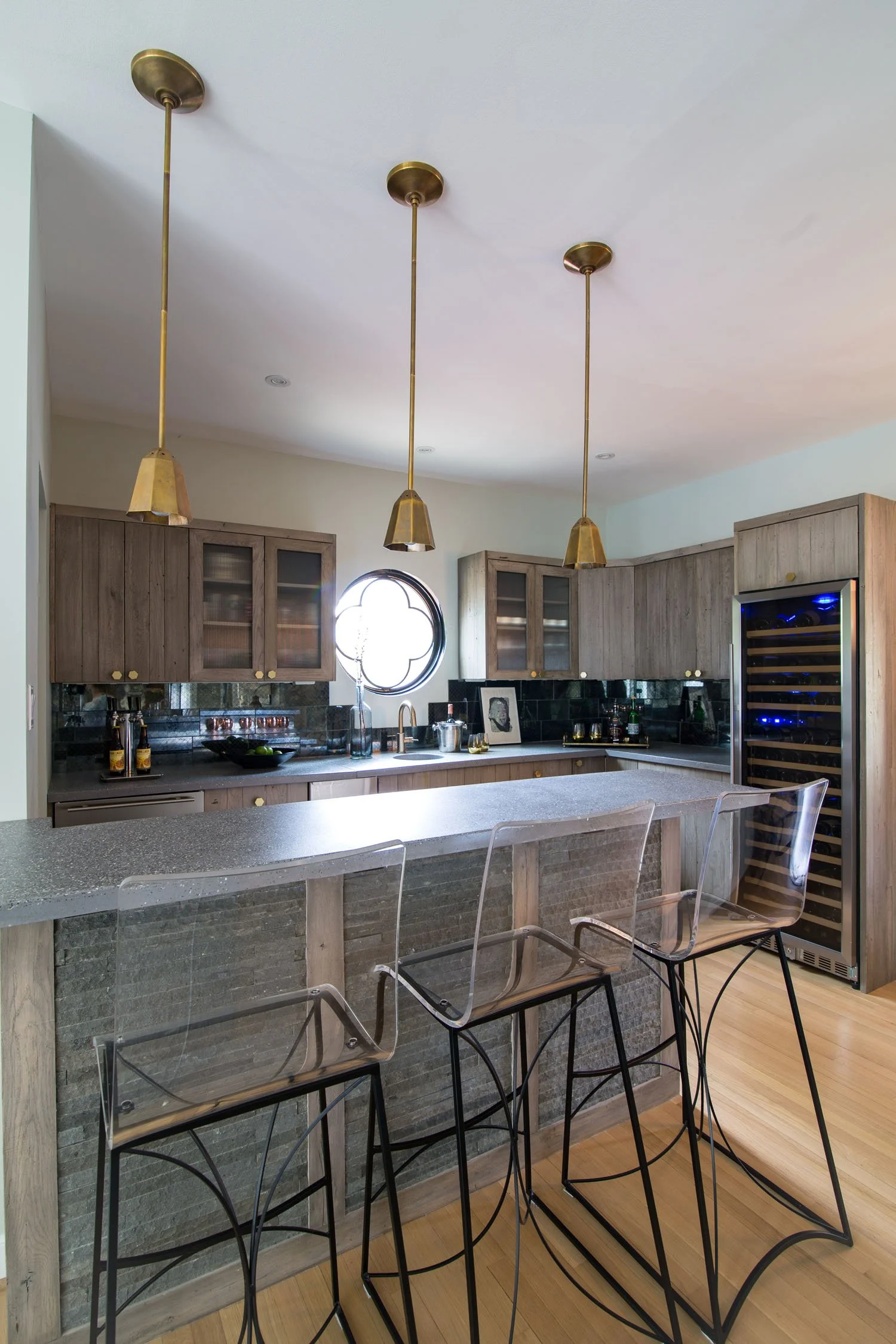 Modern kitchen with gray wood cabinets, black tiled backsplash, and pendant lights over a breakfast bar. Transparent bar stools sit at the counter, and there is a wine cooler on the right side.