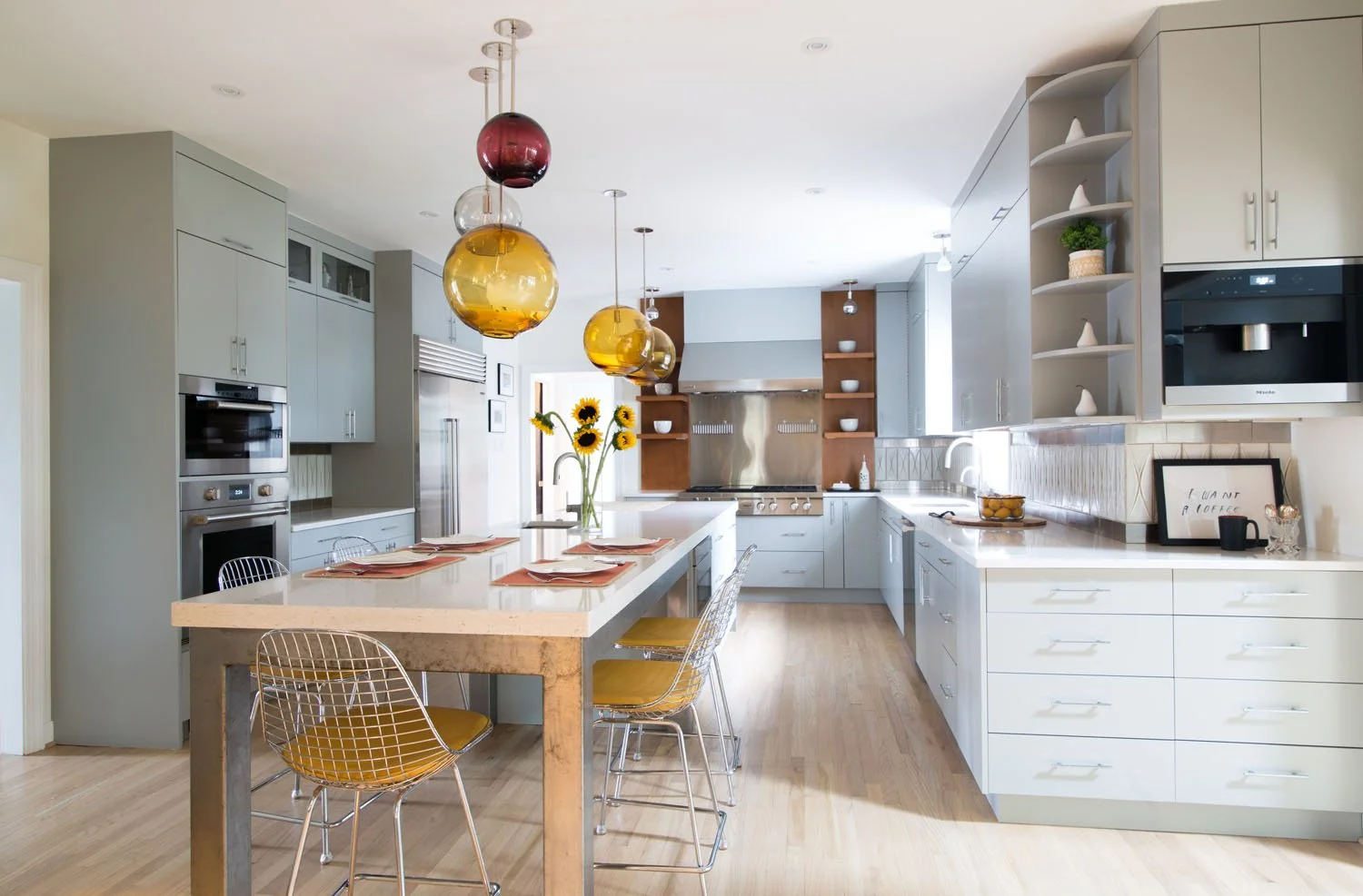 Modern kitchen with white cabinetry, stainless steel appliances, a large kitchen island with place settings and sunflowers, and colorful glass pendant lights.