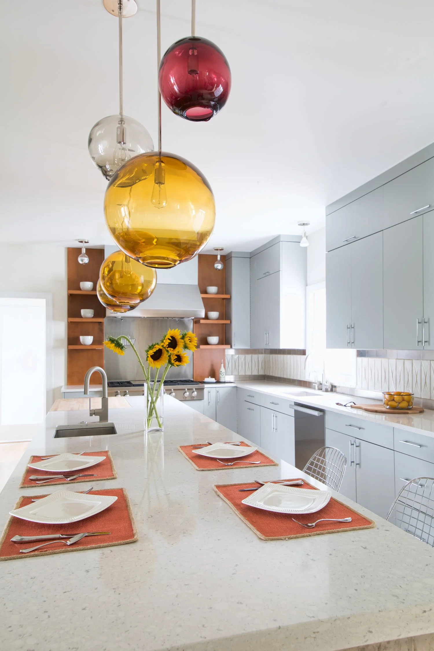 A modern kitchen with a white central island, set for four with white plates, orange placemats, and silverware, decorated with a vase of sunflowers. Hanging above are five colorful glass pendant lights in red, yellow, and clear. The background featur
