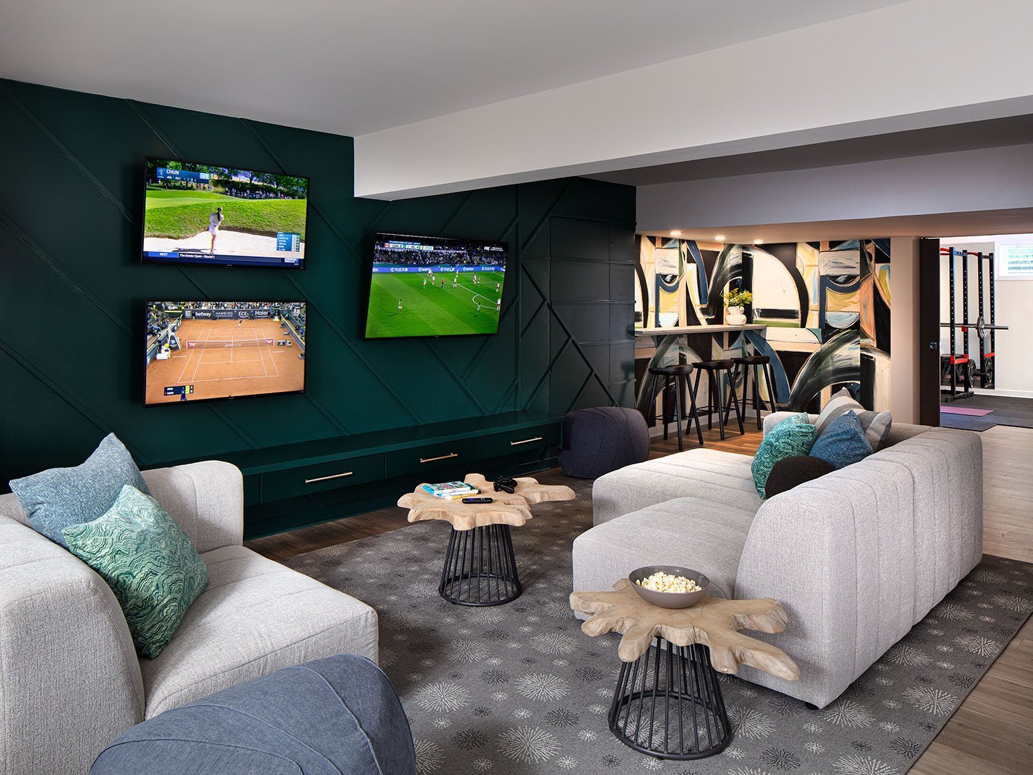 A living room with three wall-mounted TVs displaying sports games, beige sofas with colorful pillows, a gray area rug, and small wooden side tables with popcorn and remote controls. There is a bar area with stools and an abstract mural.
