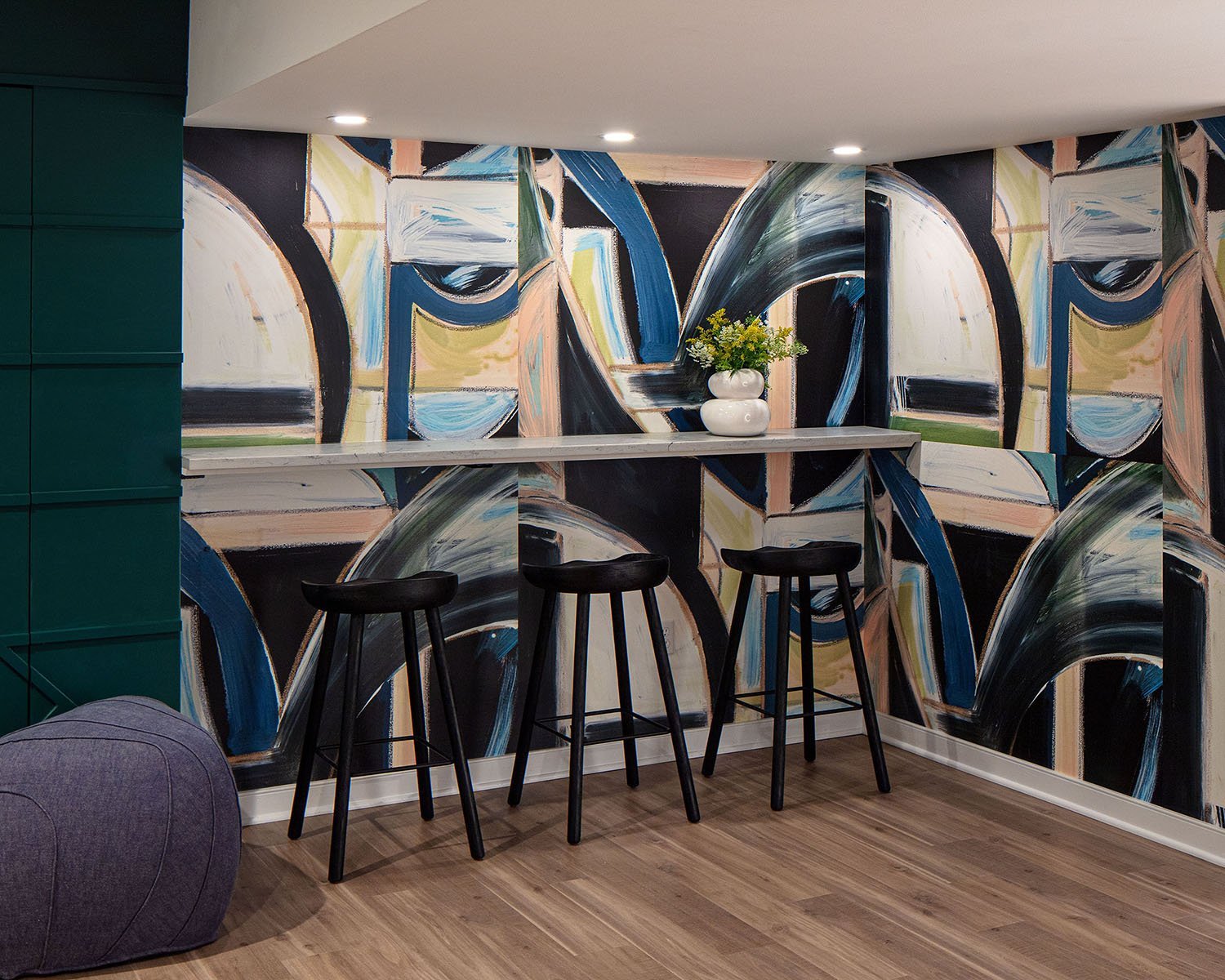 Modern interior with an abstract, colorful wall mural, a narrow white bar table with three black stools, a white vase with yellow flowers, and a purple lounge chair on a wooden floor.