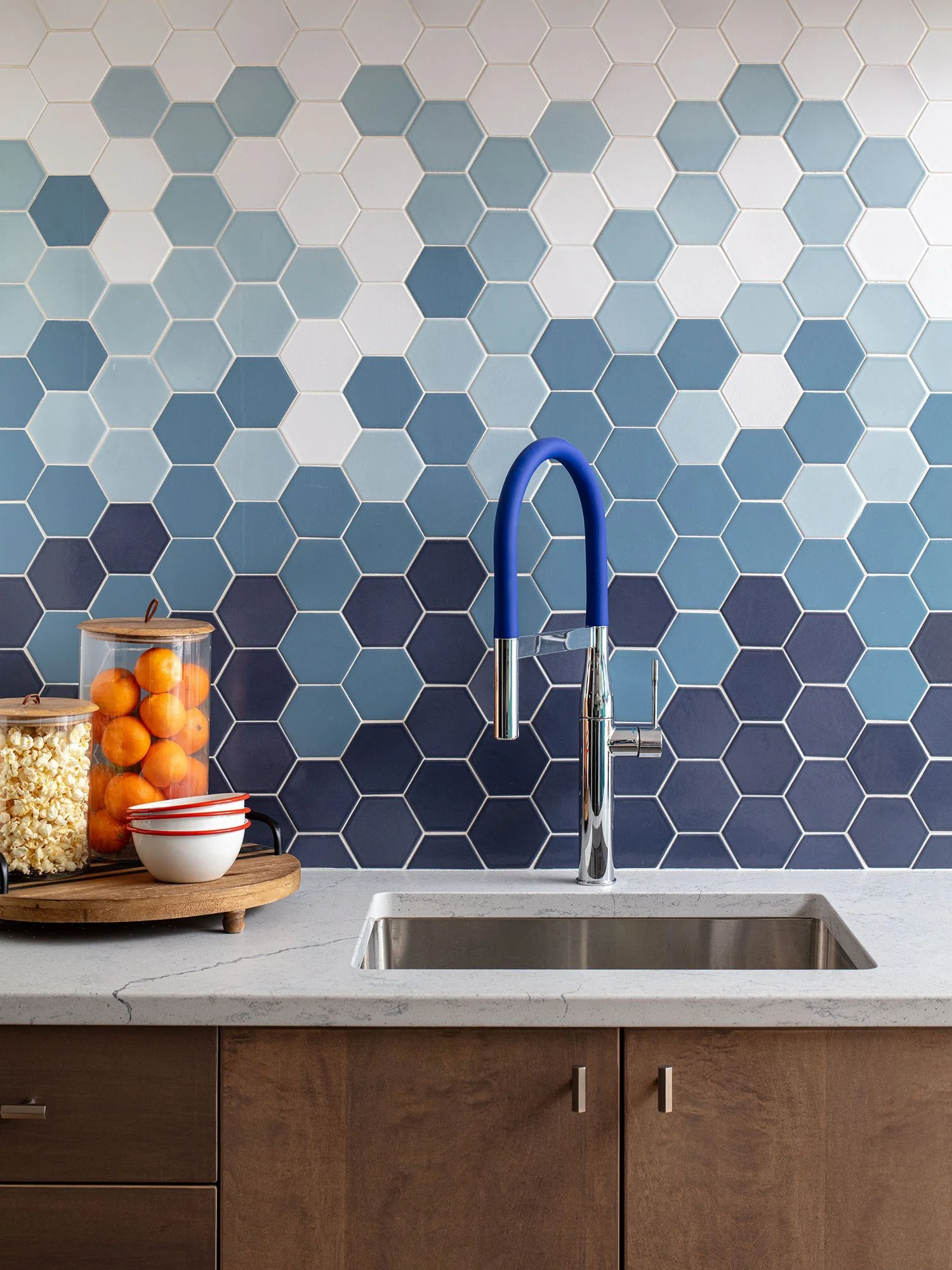 A kitchen backsplash with hexagonal tiles in various shades of blue. A stainless steel sink with a modern faucet featuring a blue hose. On the left, there are two glass jars on a wooden tray, filled with popcorn and oranges, along with a small white 