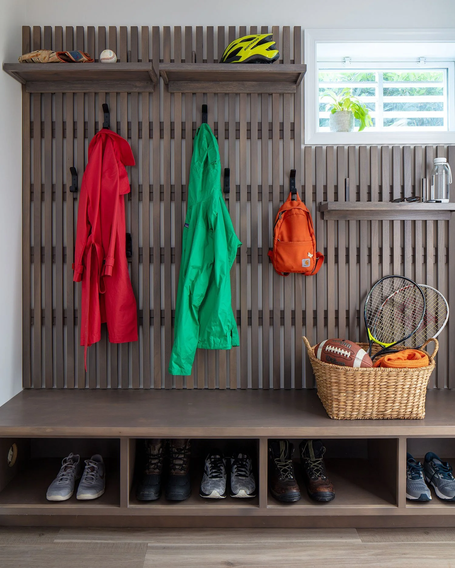Mudroom with hooks holding red, green, and orange backpacks and coats, a basket containing a football and tennis rackets, a row of shoes, and small shelves with sports gear, a small window with a potted plant.
