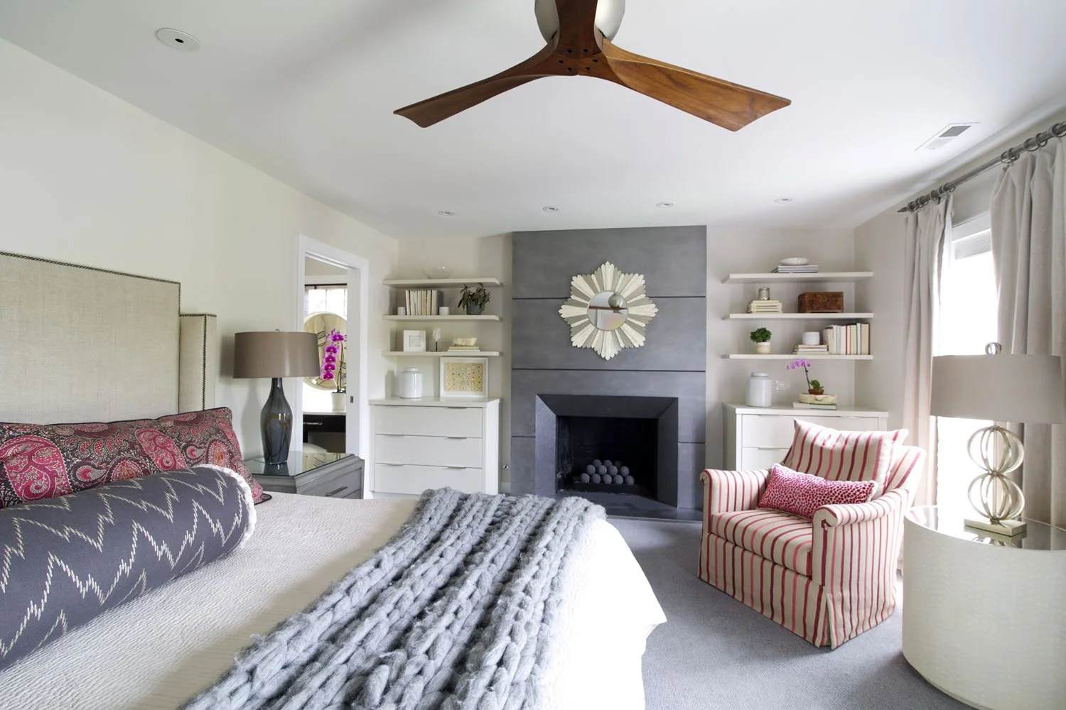 A cozy bedroom with a bed on the left, a gray and pink patterned pillow, and a chunky gray blanket. There is a bedside table with a large gray lamp. In the center against the wall is a modern fireplace with a round mirror above it. White shelves with