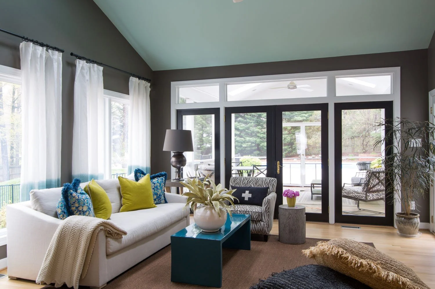 Living room with a white sofa, colorful pillows, a table with a plant, a zebra print armchair, and large glass doors leading outside to a patio with outdoor furniture and trees.