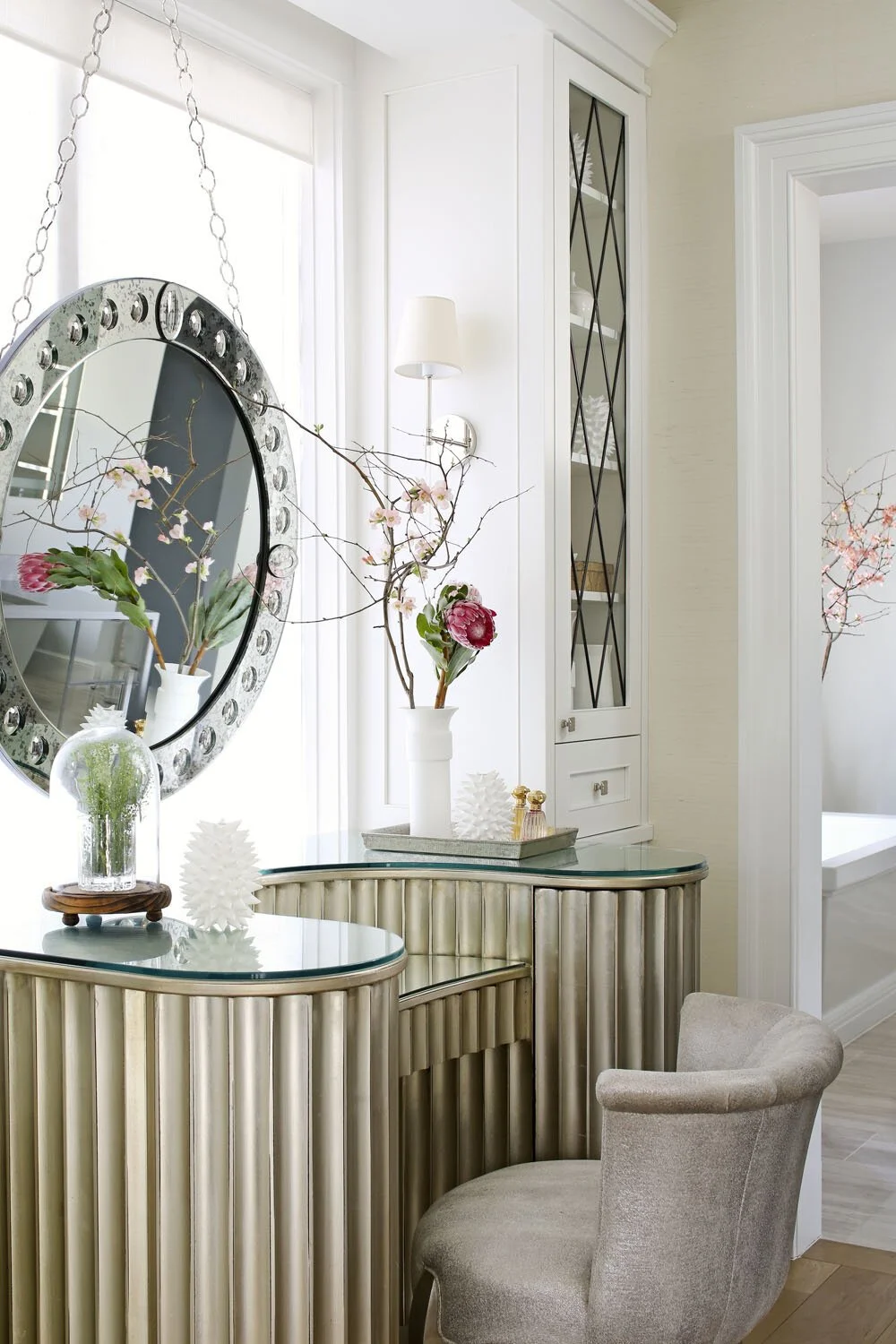 Decorative interior of a living space with a mirror, pink flowers in vases, and a gold-toned bar cabinet, styled with white and neutral tones.