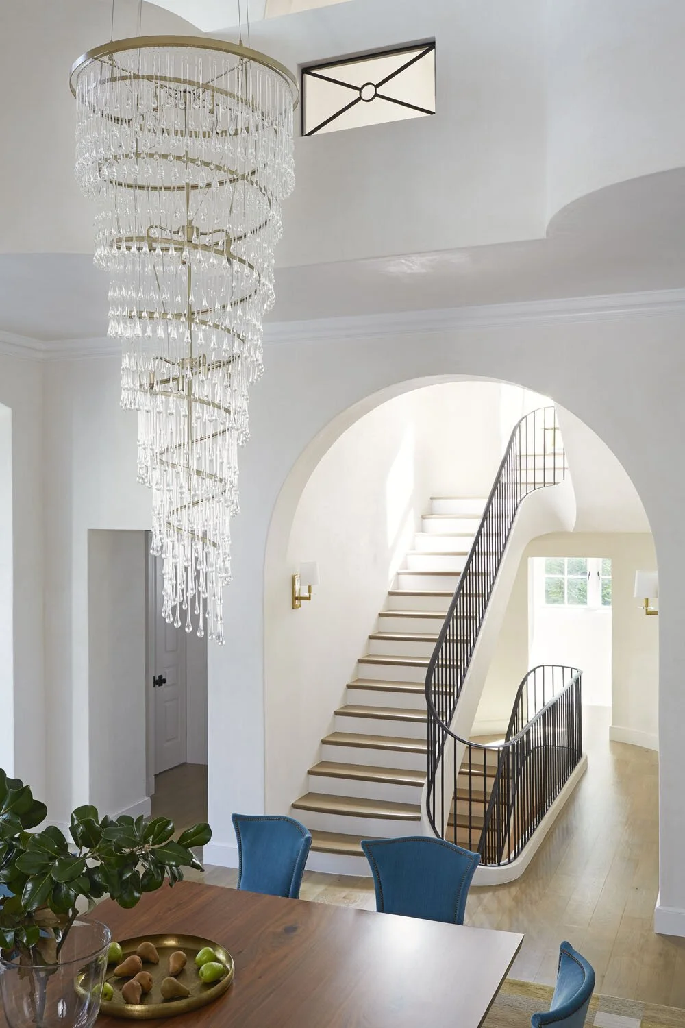 Bright interior with a spiral staircase, a large crystal chandelier hanging from the ceiling, a round wooden dining table with blue chairs, a potted plant, and a modern wall sconce.