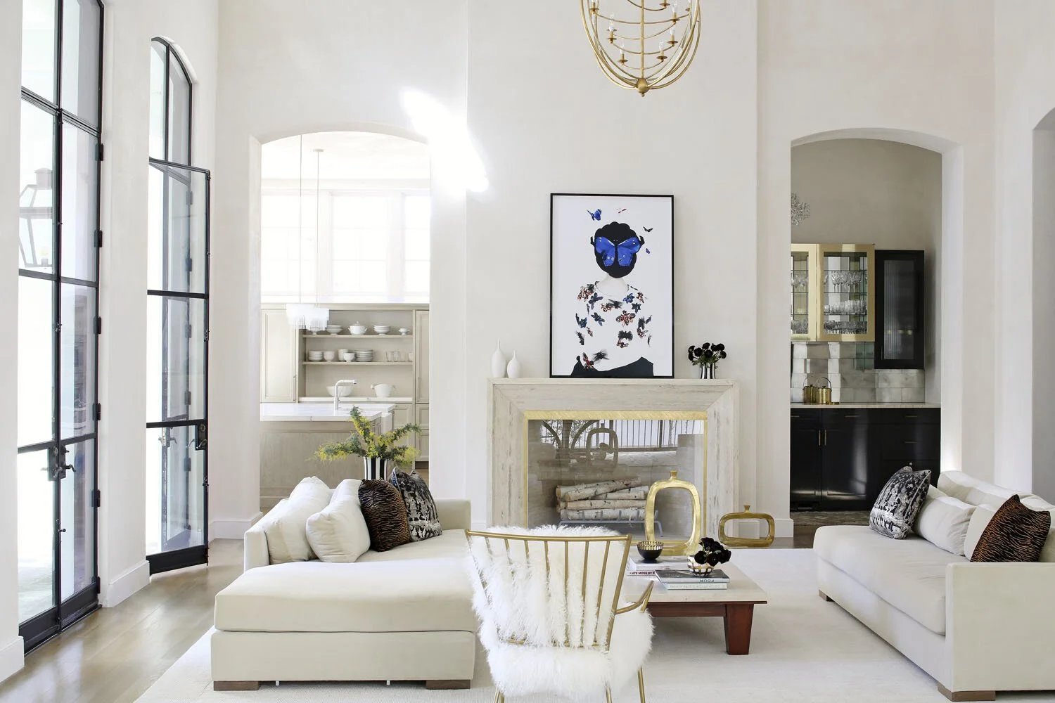 Living room with white sofas, a gold chair with a white furry cushion, a fireplace with artwork above, and a kitchen in the background.