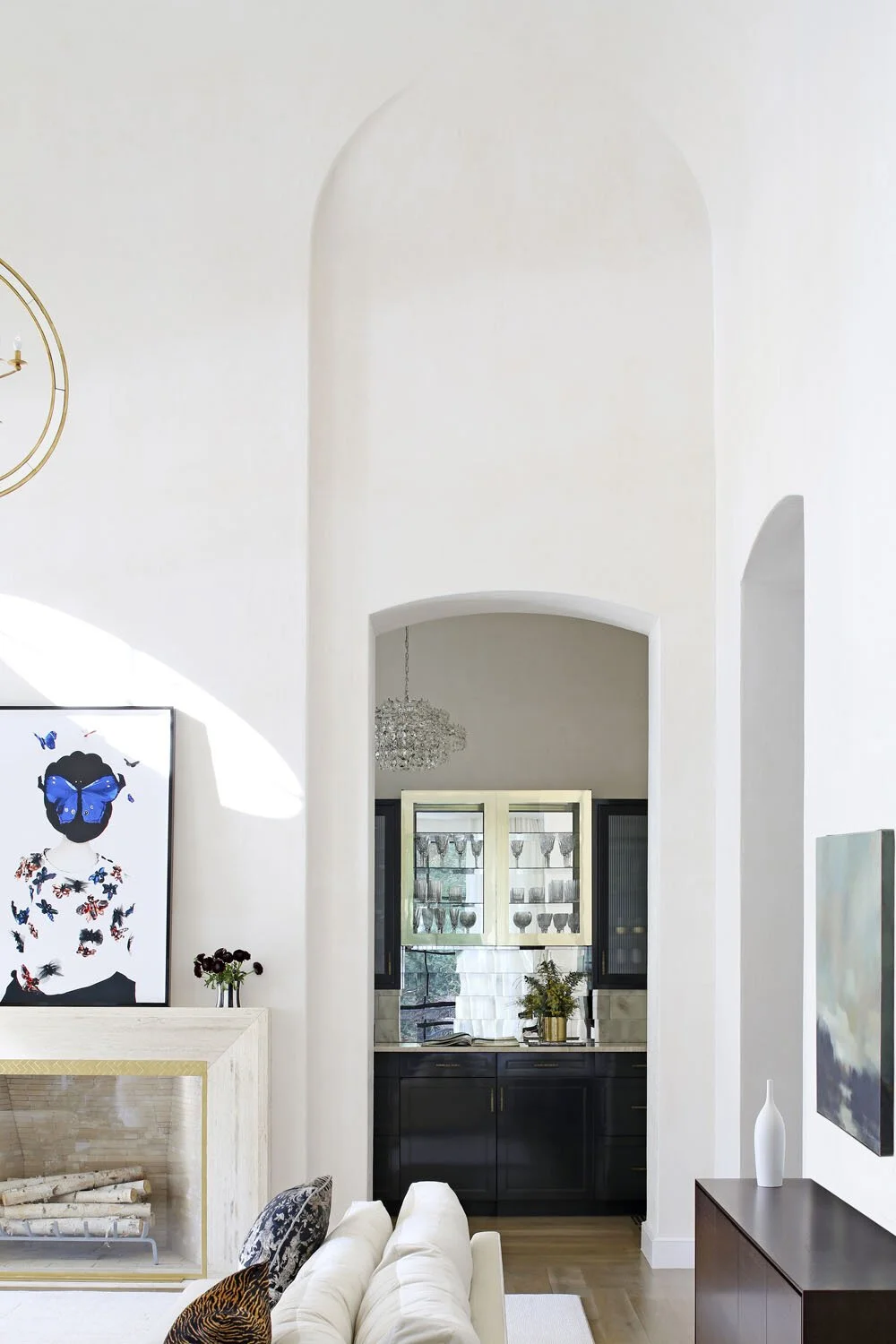 Interior view of a modern living room with white walls, a fireplace, a painting of a woman with butterfly motifs, and a dark cabinet with glassware and plants in the background.