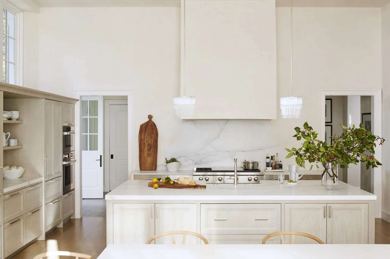 Modern kitchen with white cabinetry, marble backsplash, and a large island with a vase of greenery, hanging pendant lights, and cooking essentials on the counter.