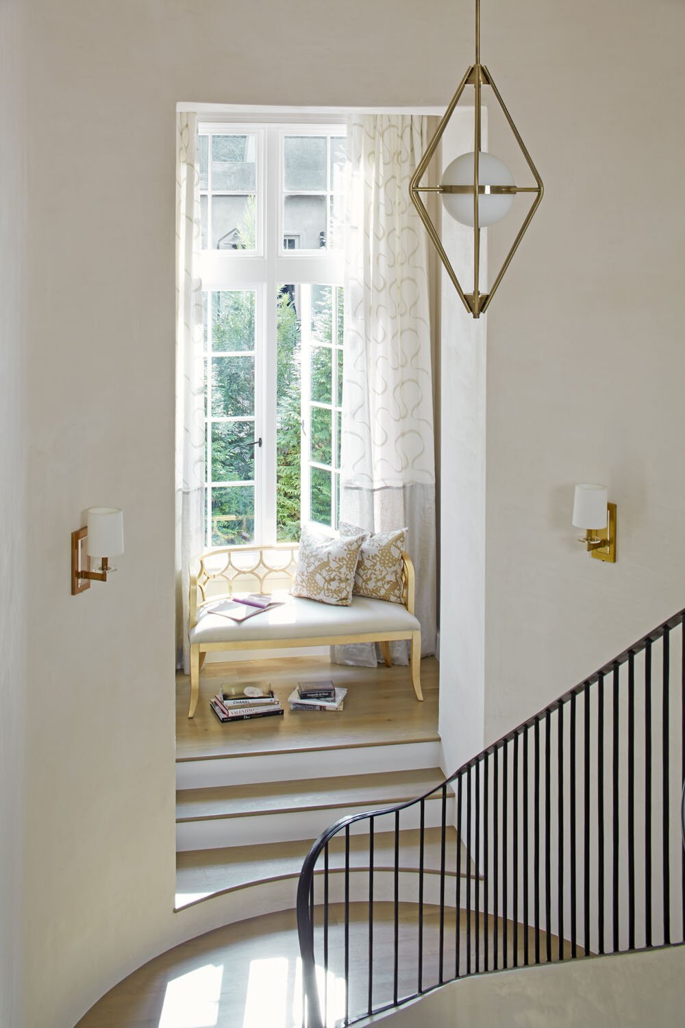 A small sitting area next to a large window with a yellow bench and decorative pillows. Books are stacked on the floor, with curtains and a modern hanging light fixture above.