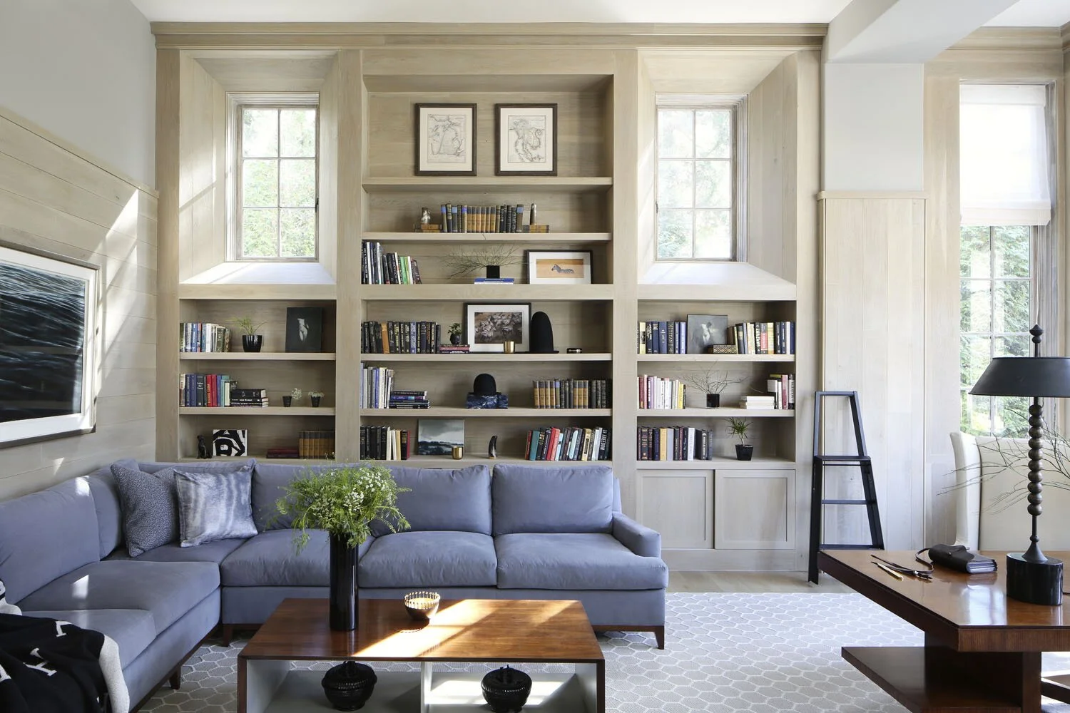 Living room with large built-in bookshelf, light-colored wood paneling, gray sofa, coffee table with plants, and windows with natural light.