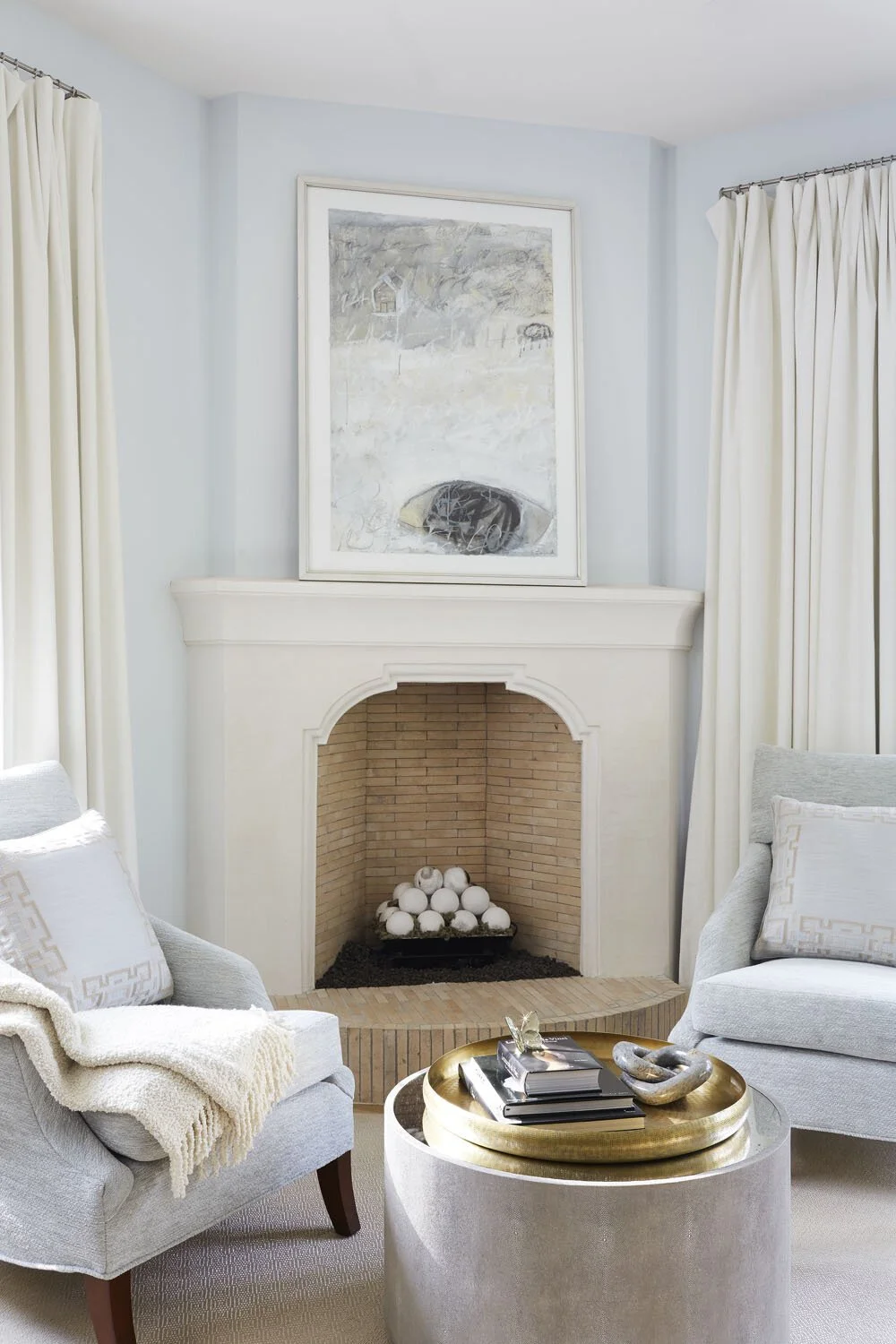 A living room corner with a white fireplace, light blue walls, white curtains, gray armchairs with pillows, and a round coffee table with books and decorative objects.